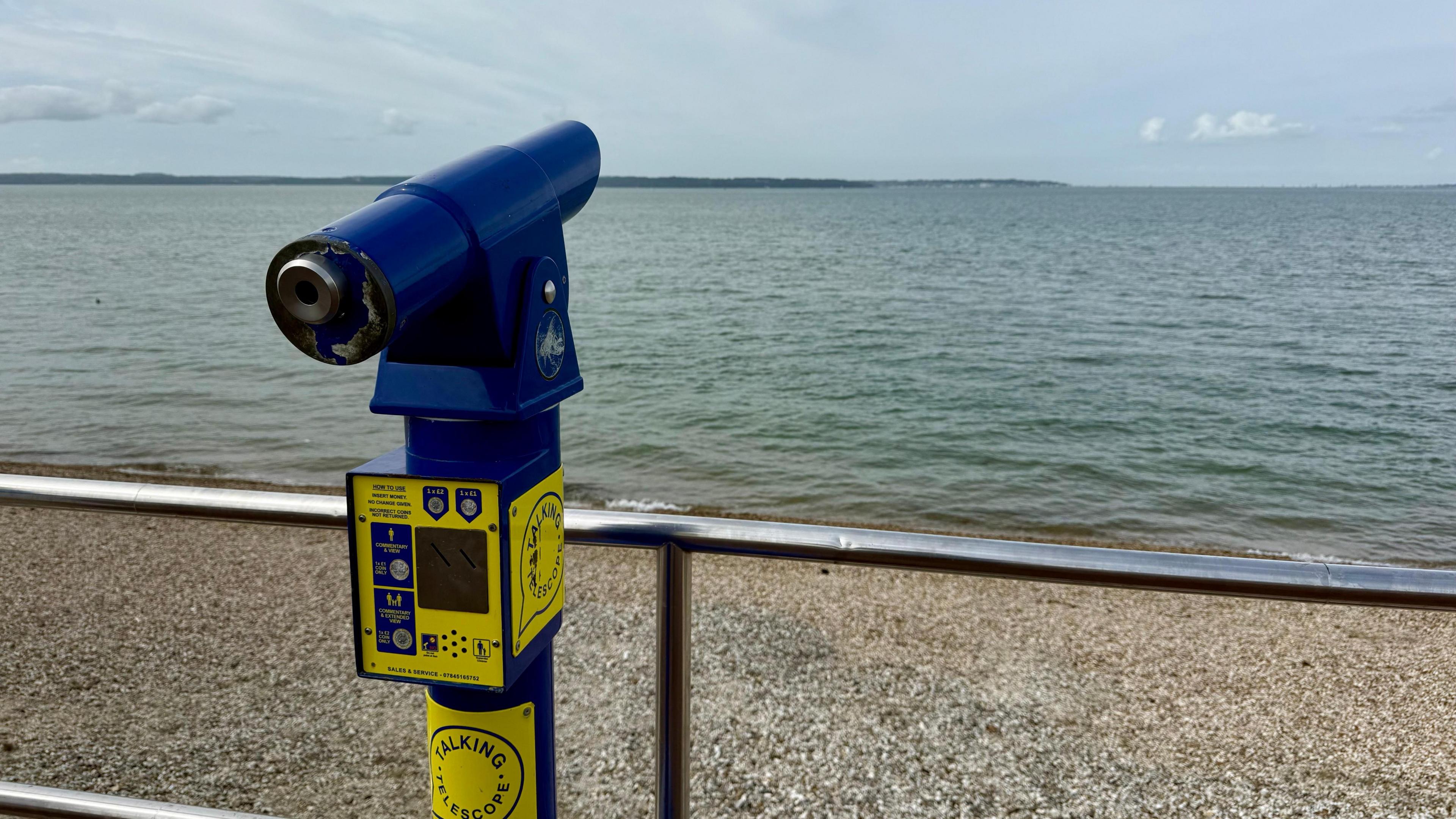 A public telescope sits on a promenade. It is dark blue with yellow stickers on it. A chrome railing is just in front of it before a pebble beach and calm seas can be seen all the way to the horizon