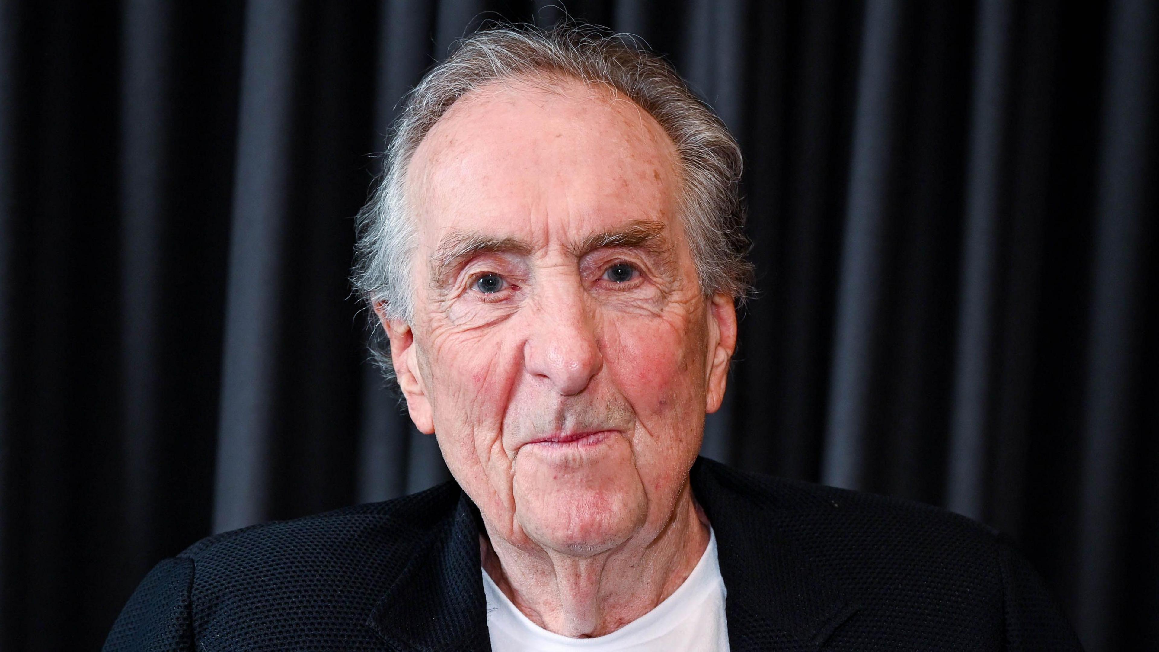 Eric Idle smiles for a photo in front of a dark curtain backdrop. He is wearing a black blazer over a white shirt.