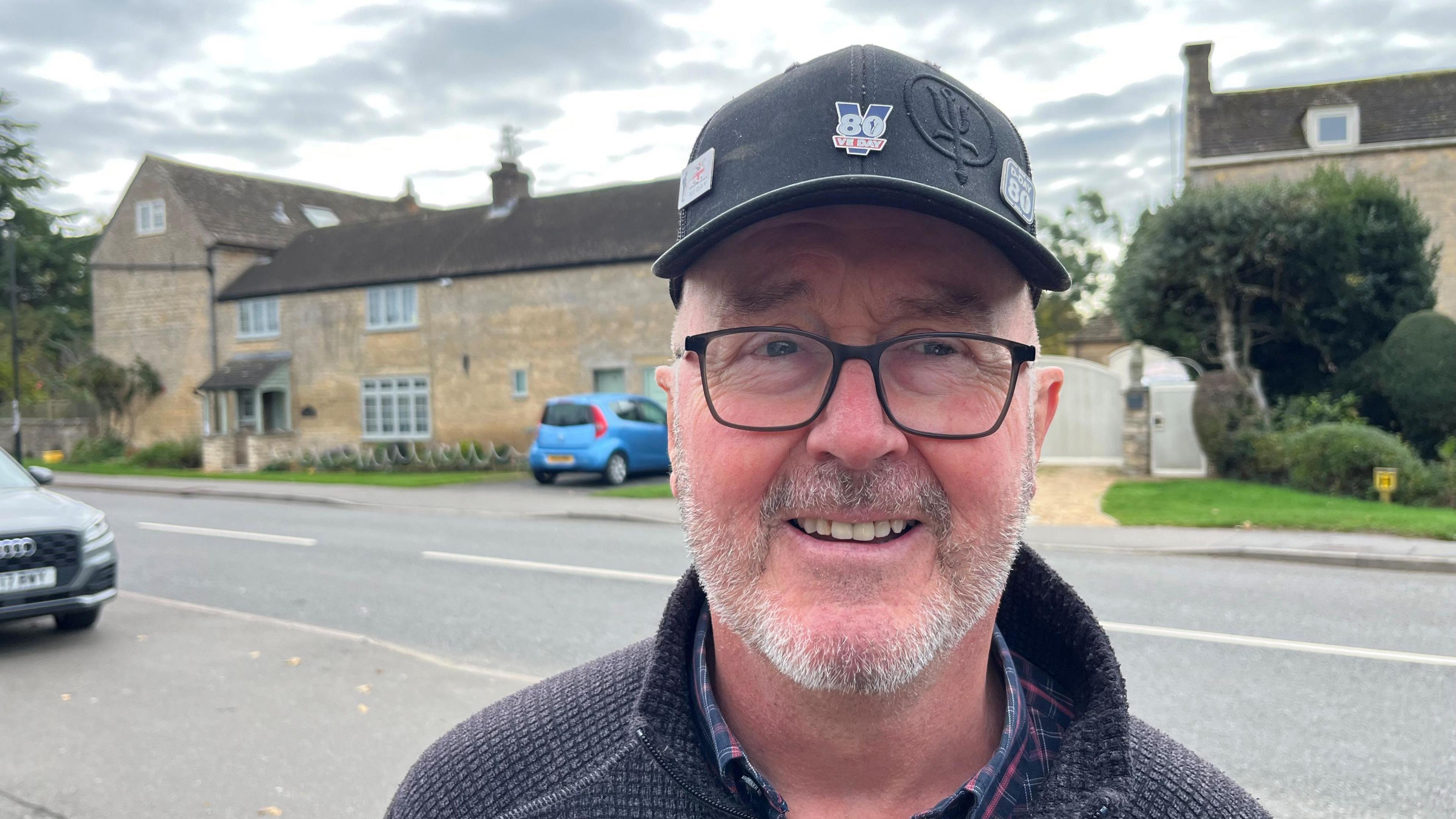 Gregg Duggan has a light brown and grey beard and moustache, with a black baseball cap on his head with some metal badges attached, he has dark framed glasses on and he is wearing a dark sweater with a checked shirt underneath. Behind him are some houses and cars parked.