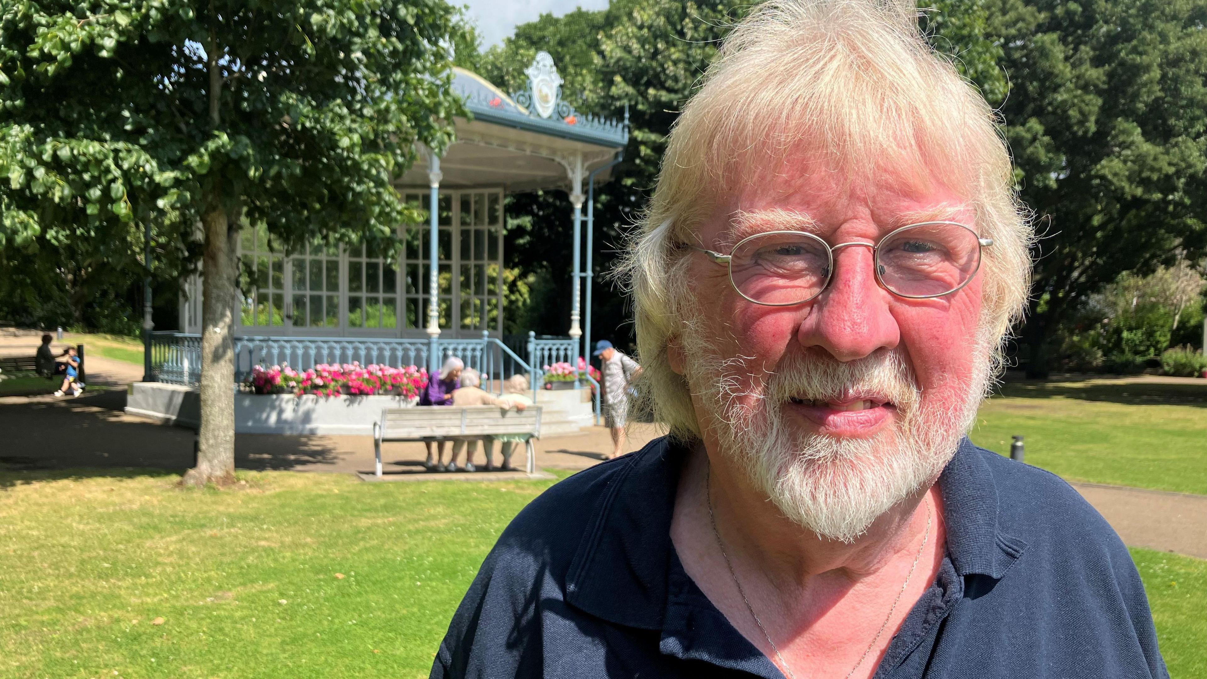 Ulster Fleadh organiser Jimi Quinn wears a navy polo shirt which has a badge on it. He has white hair, a white beard and wears wired-rimmed glasses. He is stood outside in front of a bandstand.