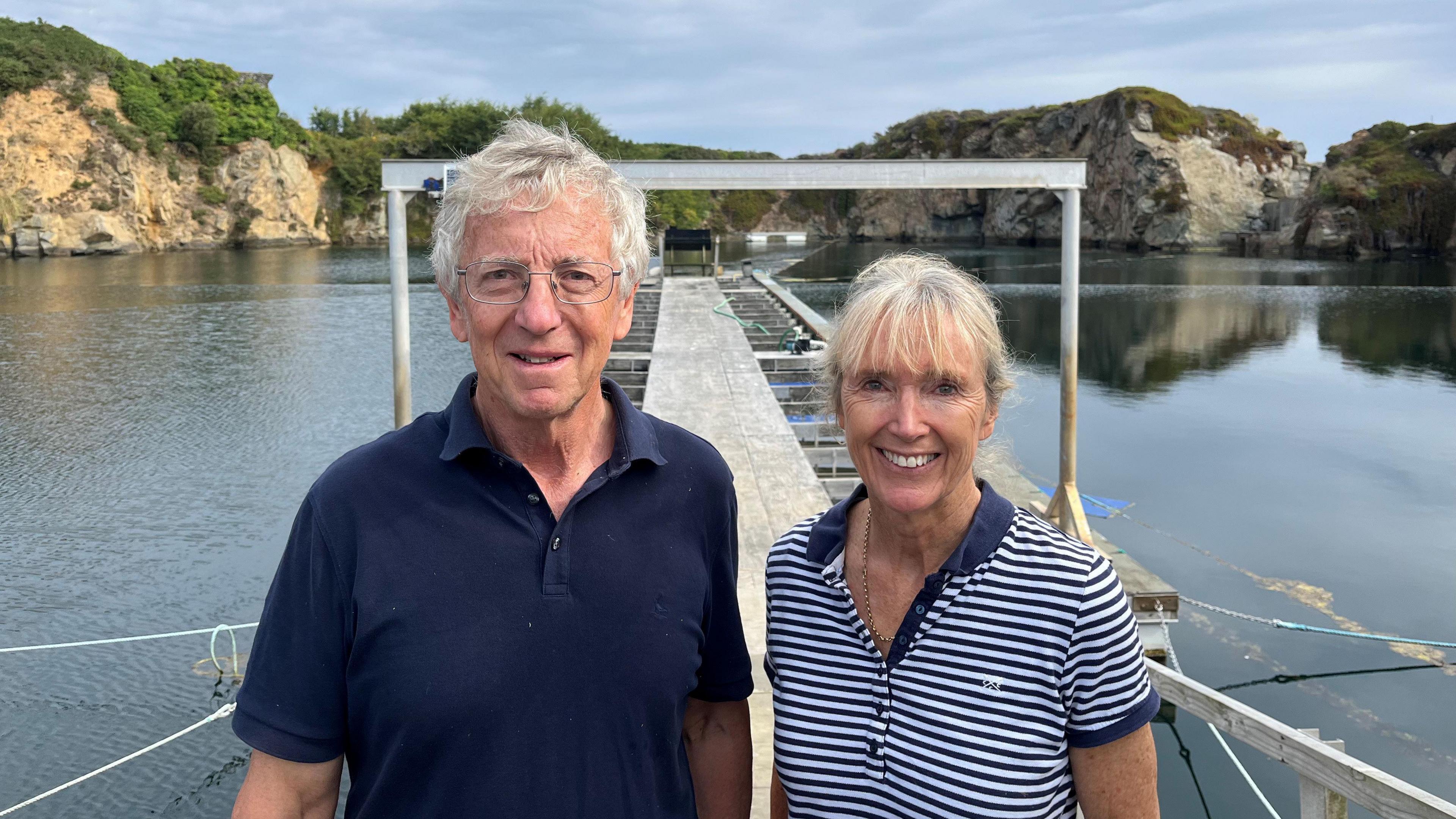 A man with grey hair and glasses in a blue polo shirt stands next to a grey haired woman in a blue and white hooped top. They stand in front of a jetty in a flooded former quarry.