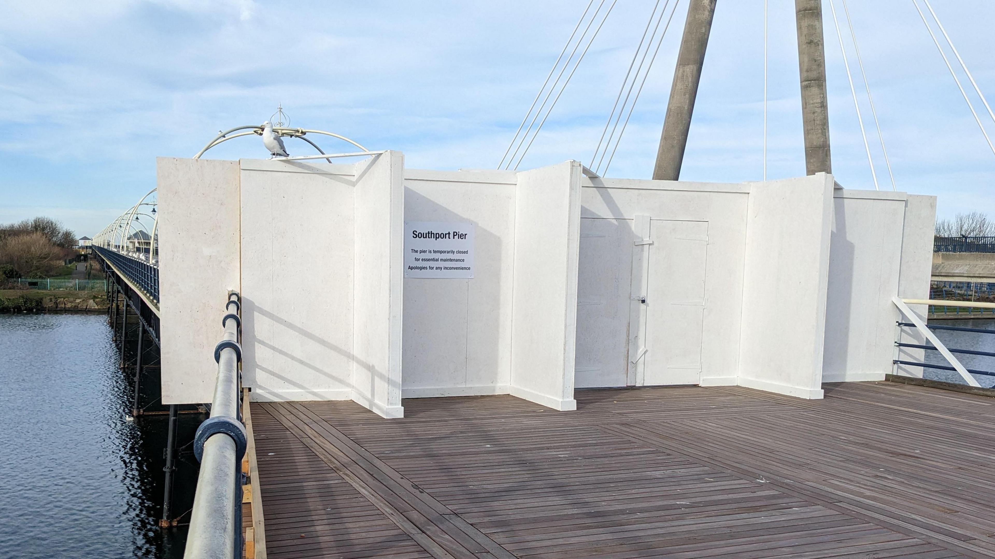 The wooden decked entrance to Southport Pier is boarded off with white boards and a sign saying the pier is closed. In the background you can see the length of the pier, surrounded by water.