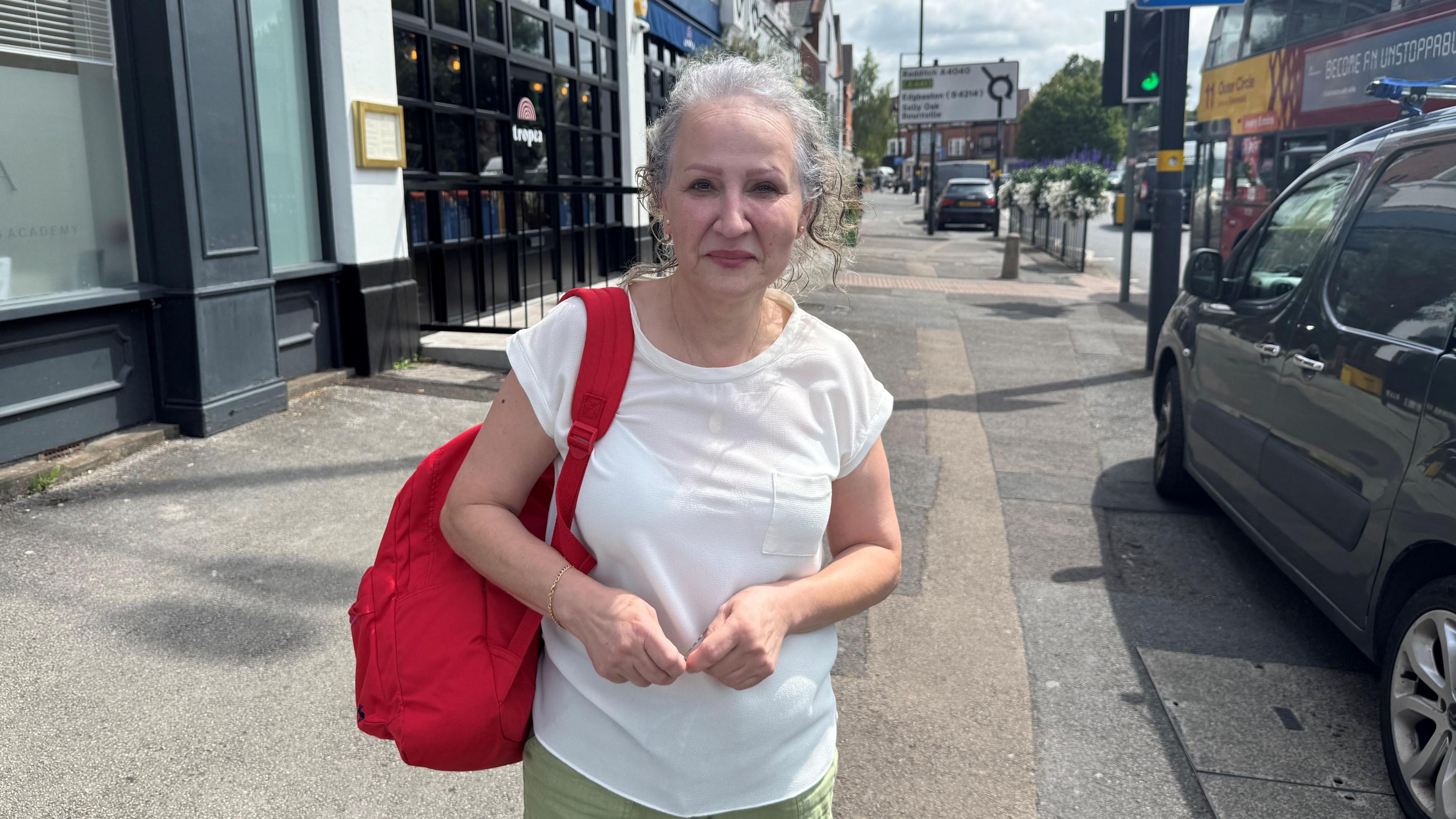 Mara Assad in a picture on the busy Harborne high street. There is a bus and a car and signs behind her. She has her hands in front of her. She has grey hair and is wearing a red backpack, a white top and green trousers.