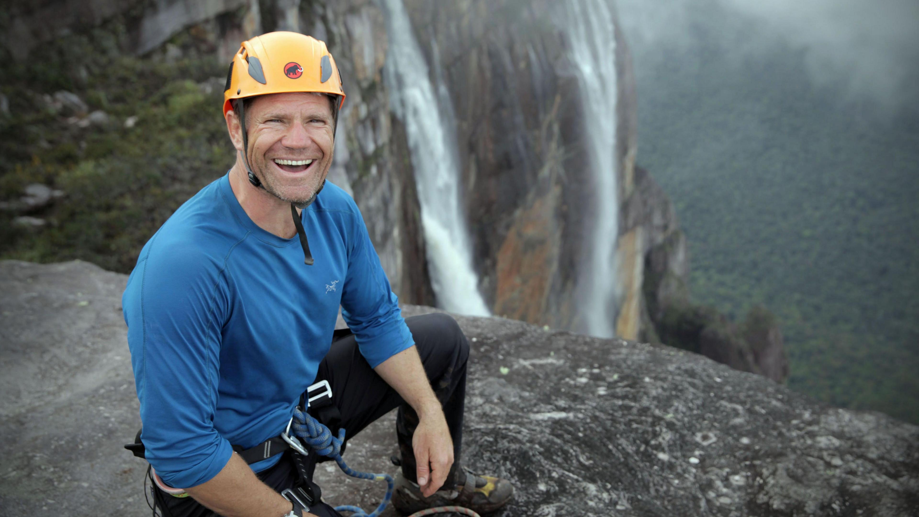 Steve Backshall wears a blue top and a yellow helment. He is kneeling on a mountain with a waterfall and forest canopy below him in the background