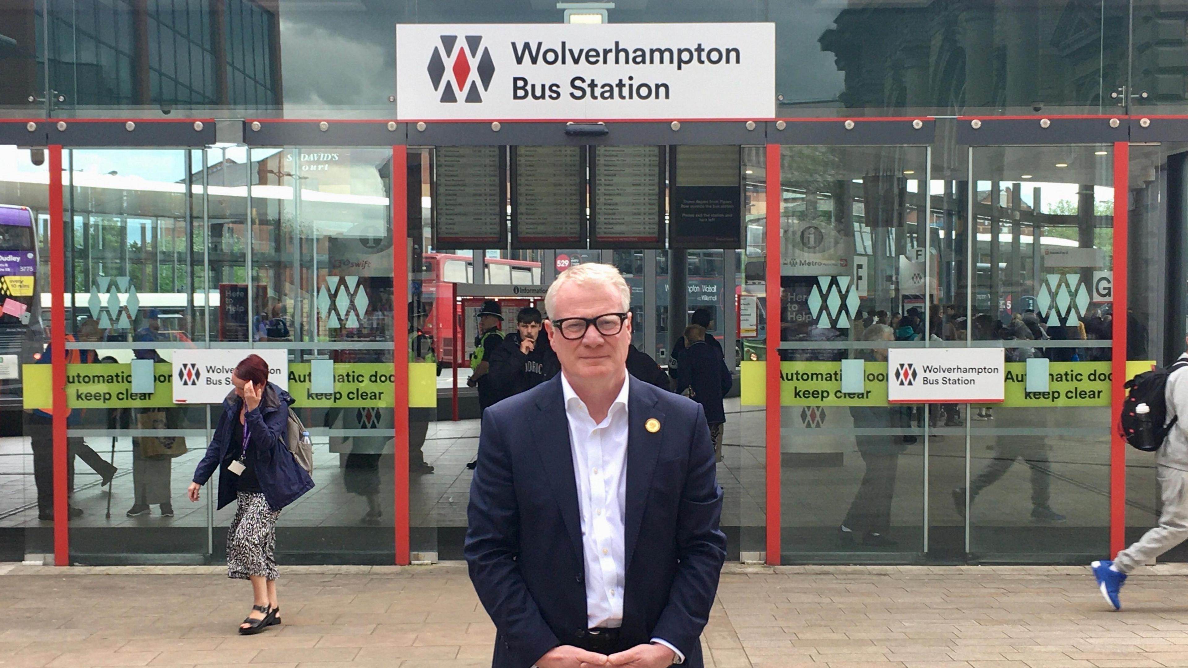 The West Midlands mayor Richard Parker, a middle-aged man with white hair and spectacles, stands in front of a set of glass doors under a sign reading "Wolverhampton Bus Station". He is wearing a blue suit jacket and a white open-necked shirt.