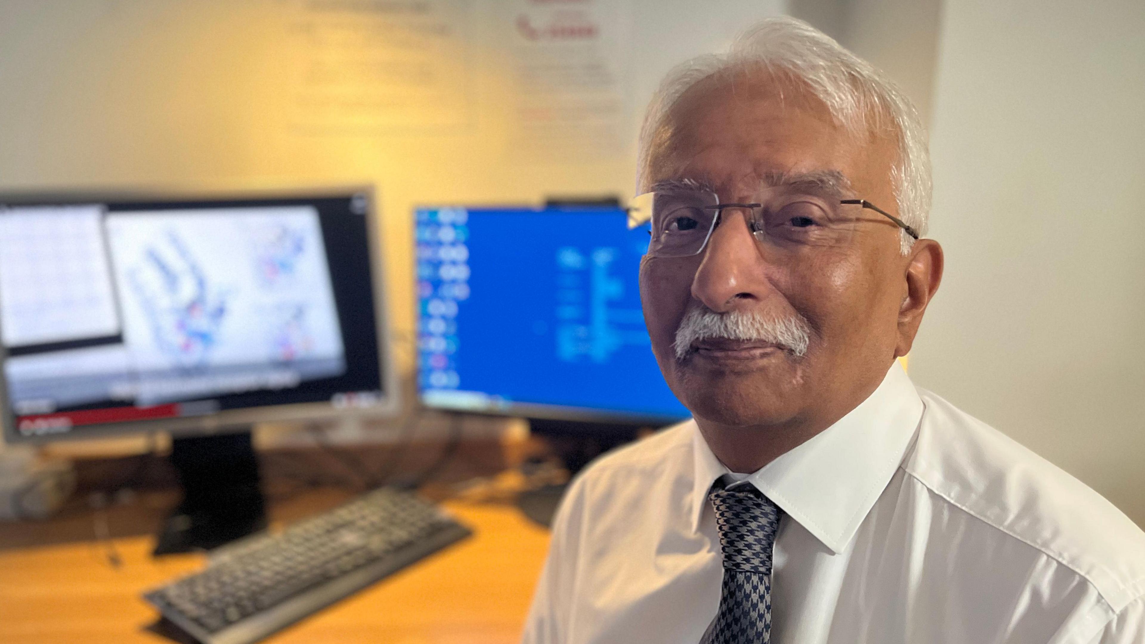 Krishna Chatterjee, with short white hair and a white moustache and wearing frameless glasses and a white shirt with a blue tie. He is smiling at the camera and behind him is a desk with two monitors and a keyboard.