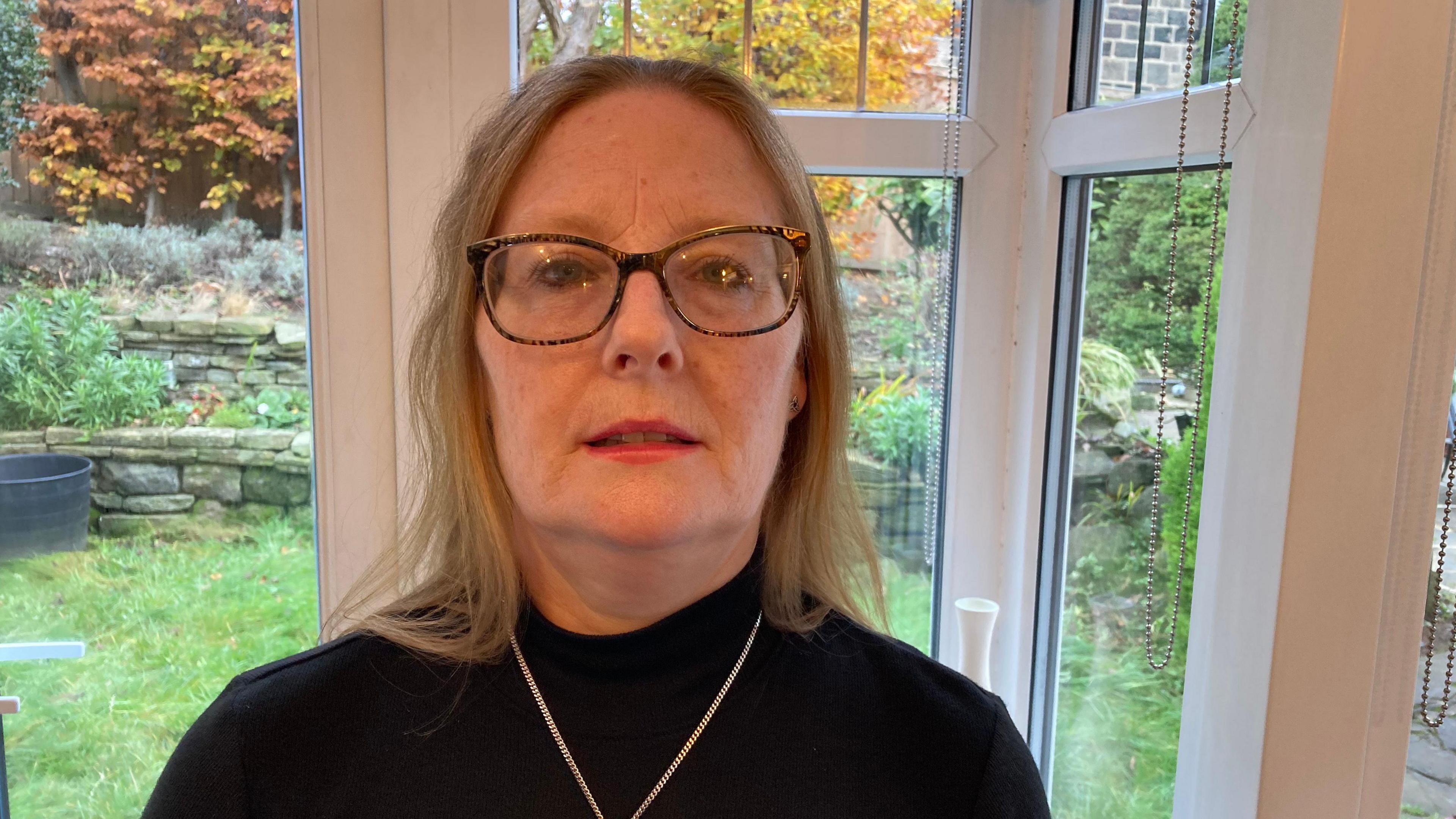 Pauline Jones - A middle-aged fair-haired lady, wearing a black top, necklace and glasses.