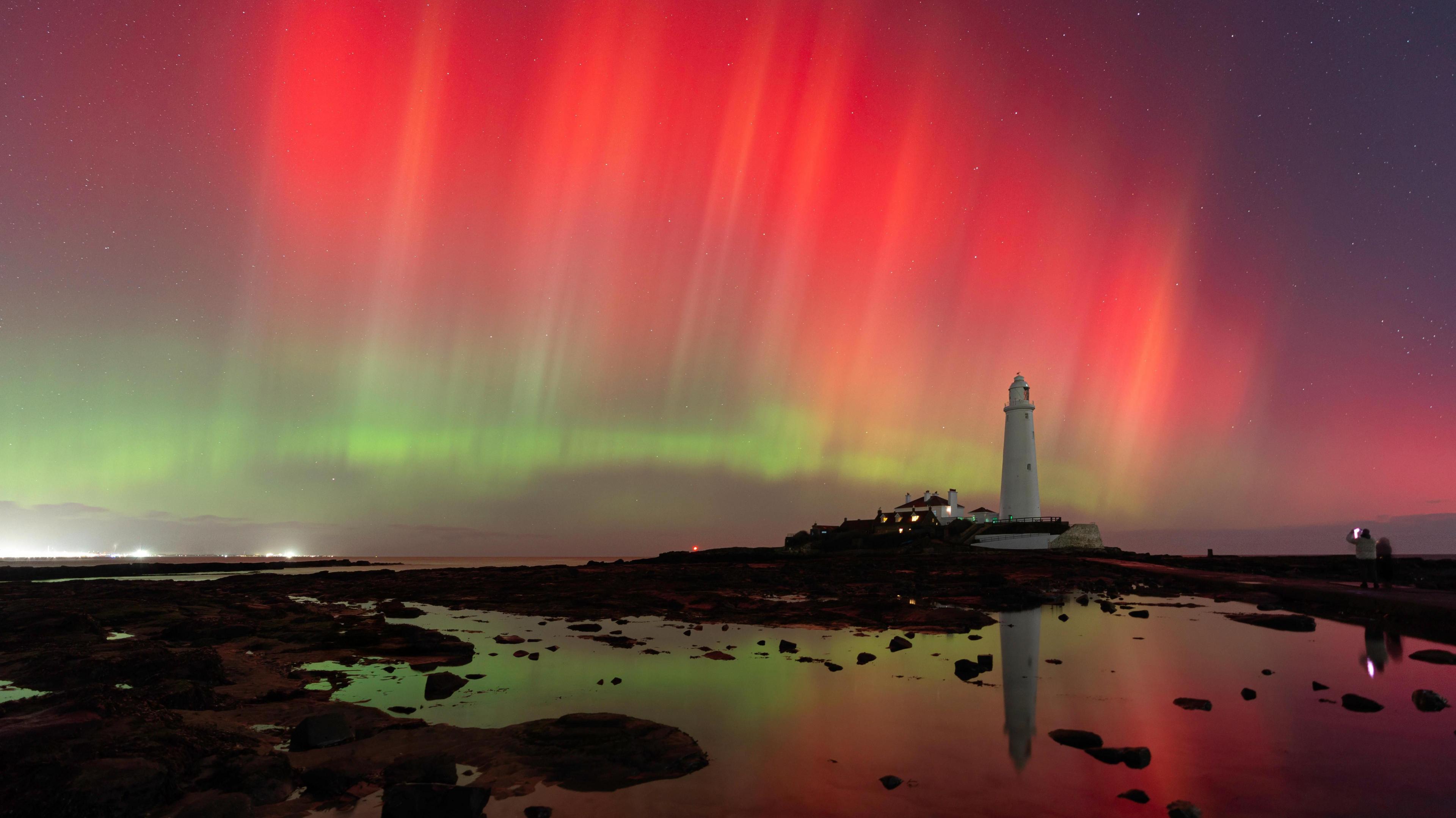 A white lighthouse with a blaze of red and green lights in the sky behind it, all reflected in a pool of water in front