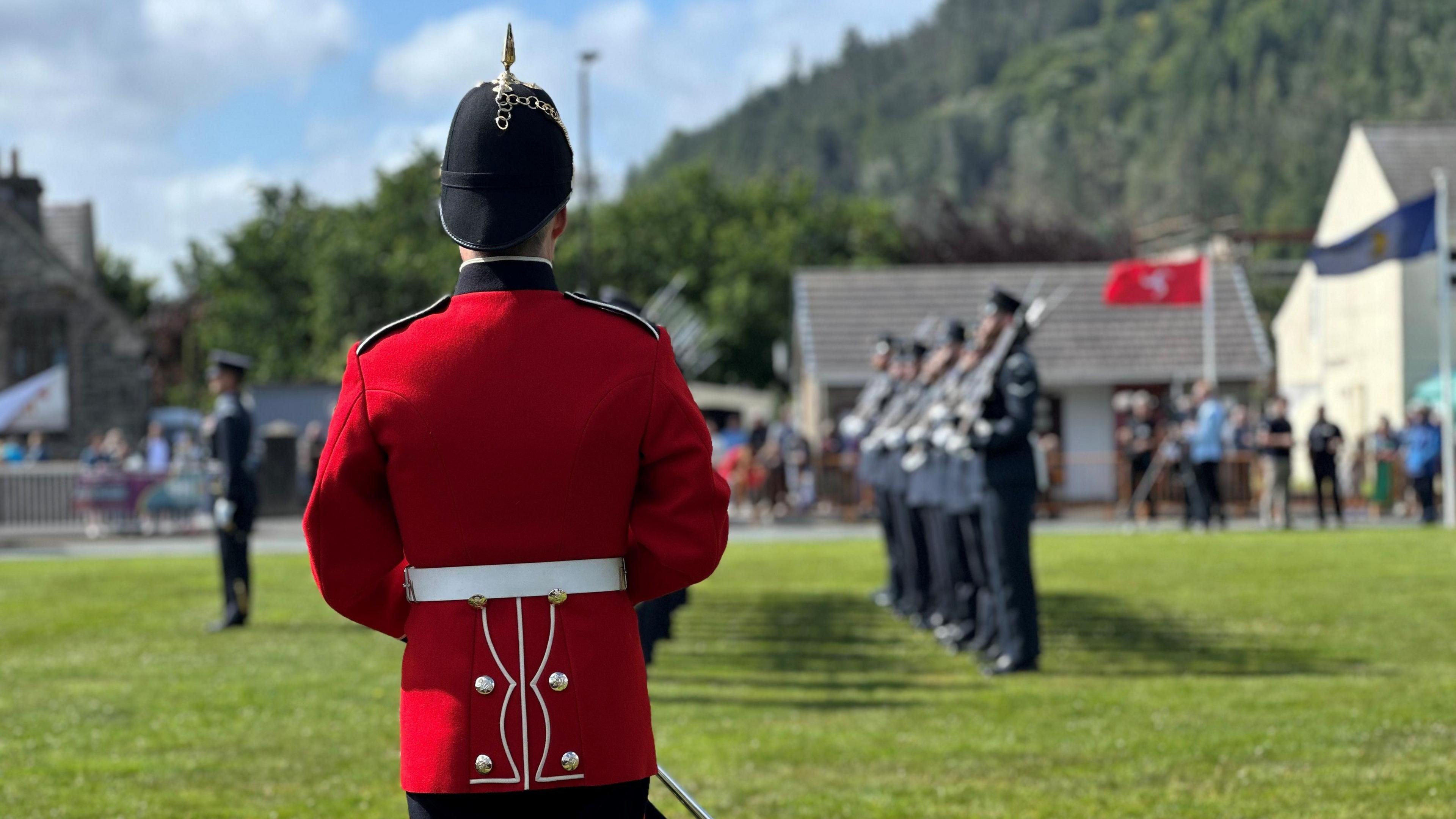 The back of a trumpeter dressed in red, who is facing the Guard of Honour, which is lined up on the green in front of him.