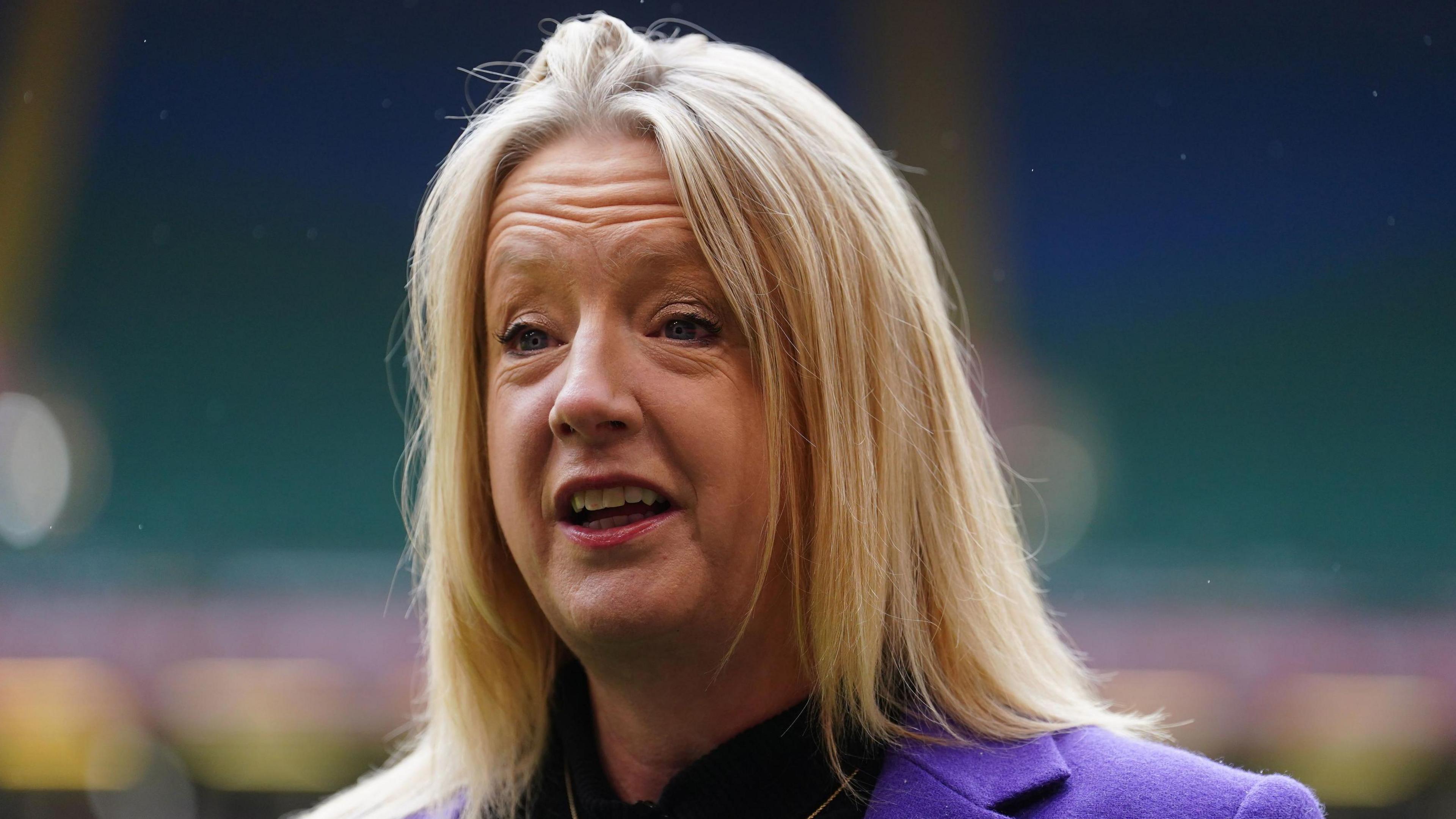 A head and shoulders picture of Abi Tierney smiling and looking to the side of the camera. She is wearing a light purple jacket over a dark top. Stands in the Principality Stadium can be seen blurred in the background.