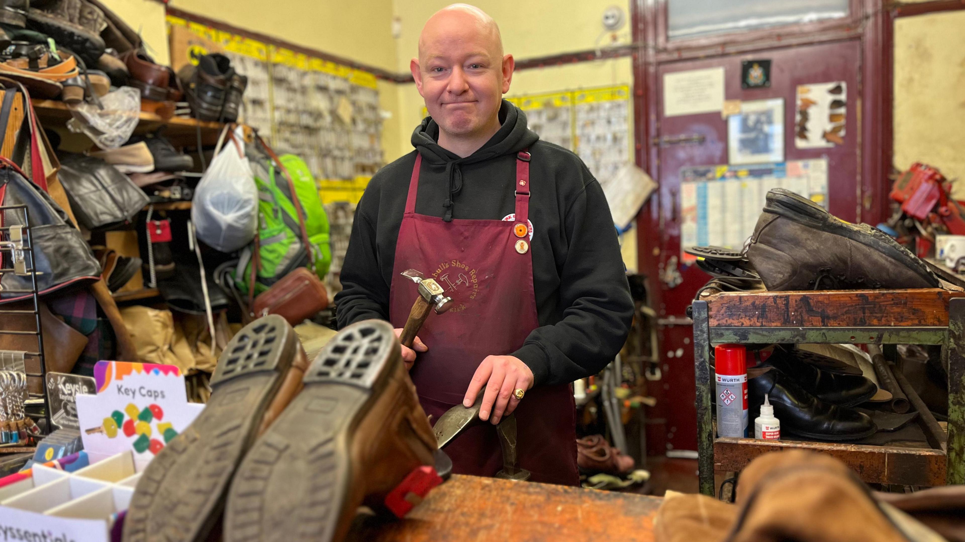 A bald-headed man in a black top with a wine-coloured apron holding a hammer above a pair of boots in a clustered workshop.