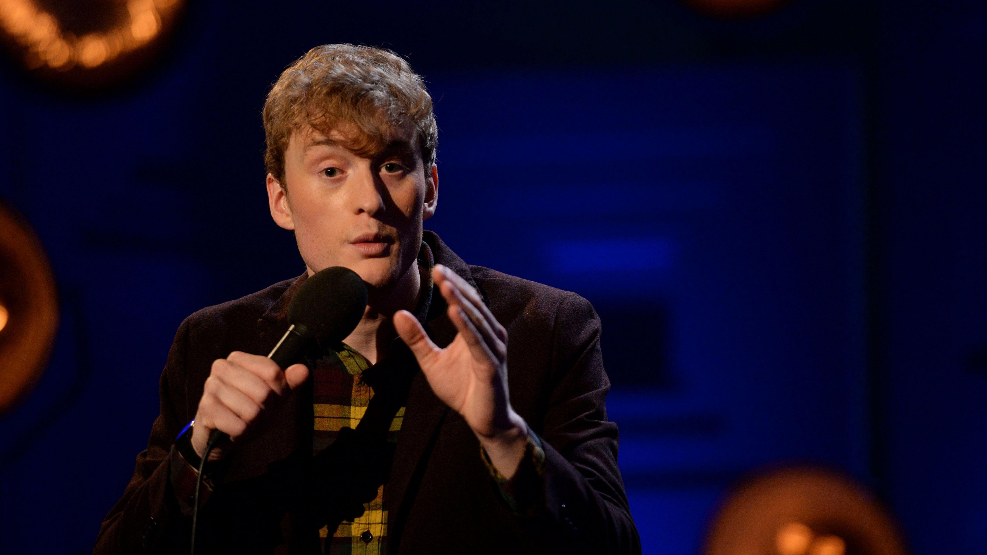 James Acaster holding a microphone delivering a stand-up comedy set