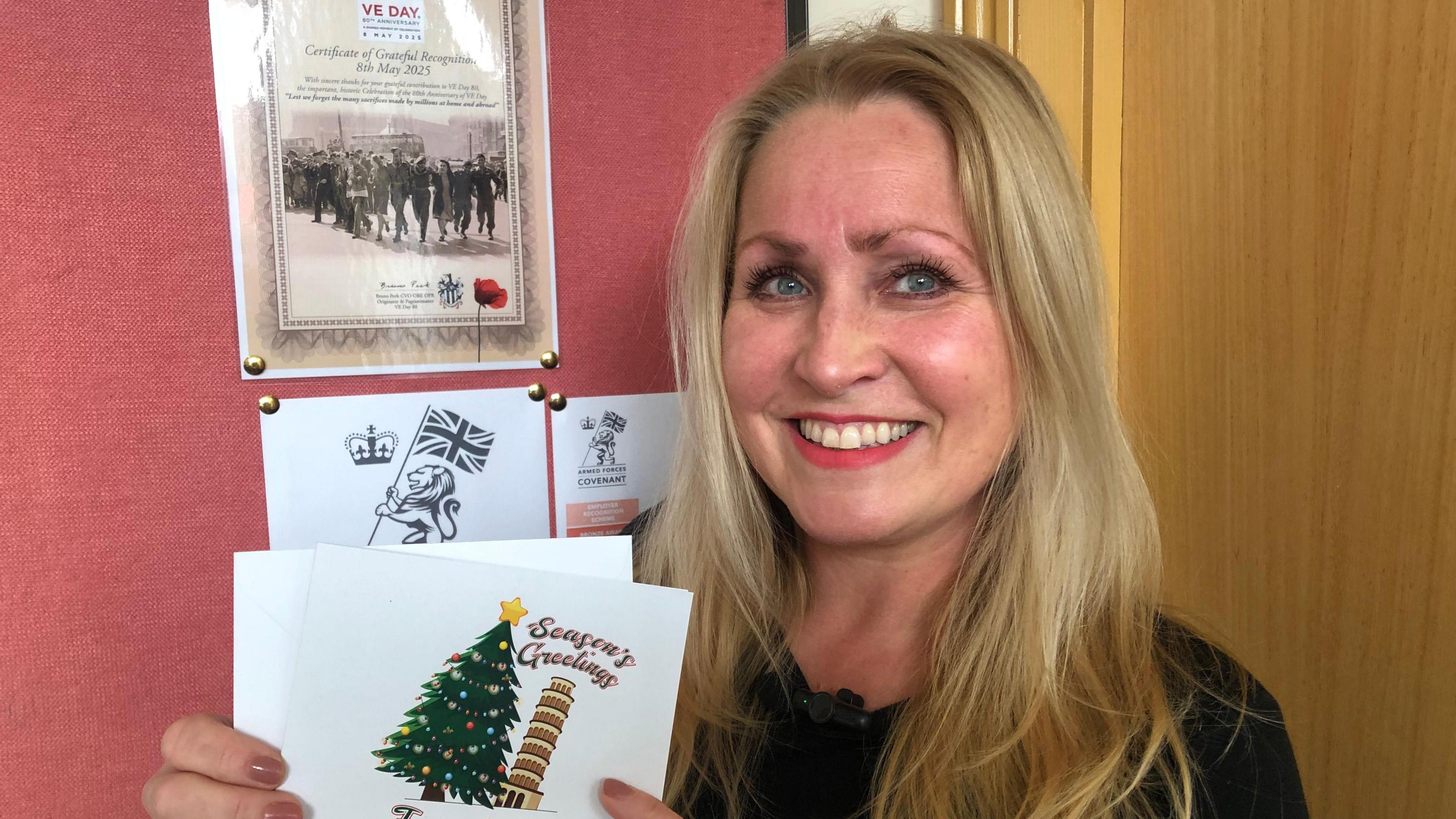 Sarah Lemmon, 50, in the March Town Council office with a notice board behind her with various posters on. She has long blonde hair, with a dark round collared jumper and is smiling, holding a greetings card from last year that shows a wonky tree next to the Leaning Tower of Pisa, with the caption "Seasons Greetings" at the top.