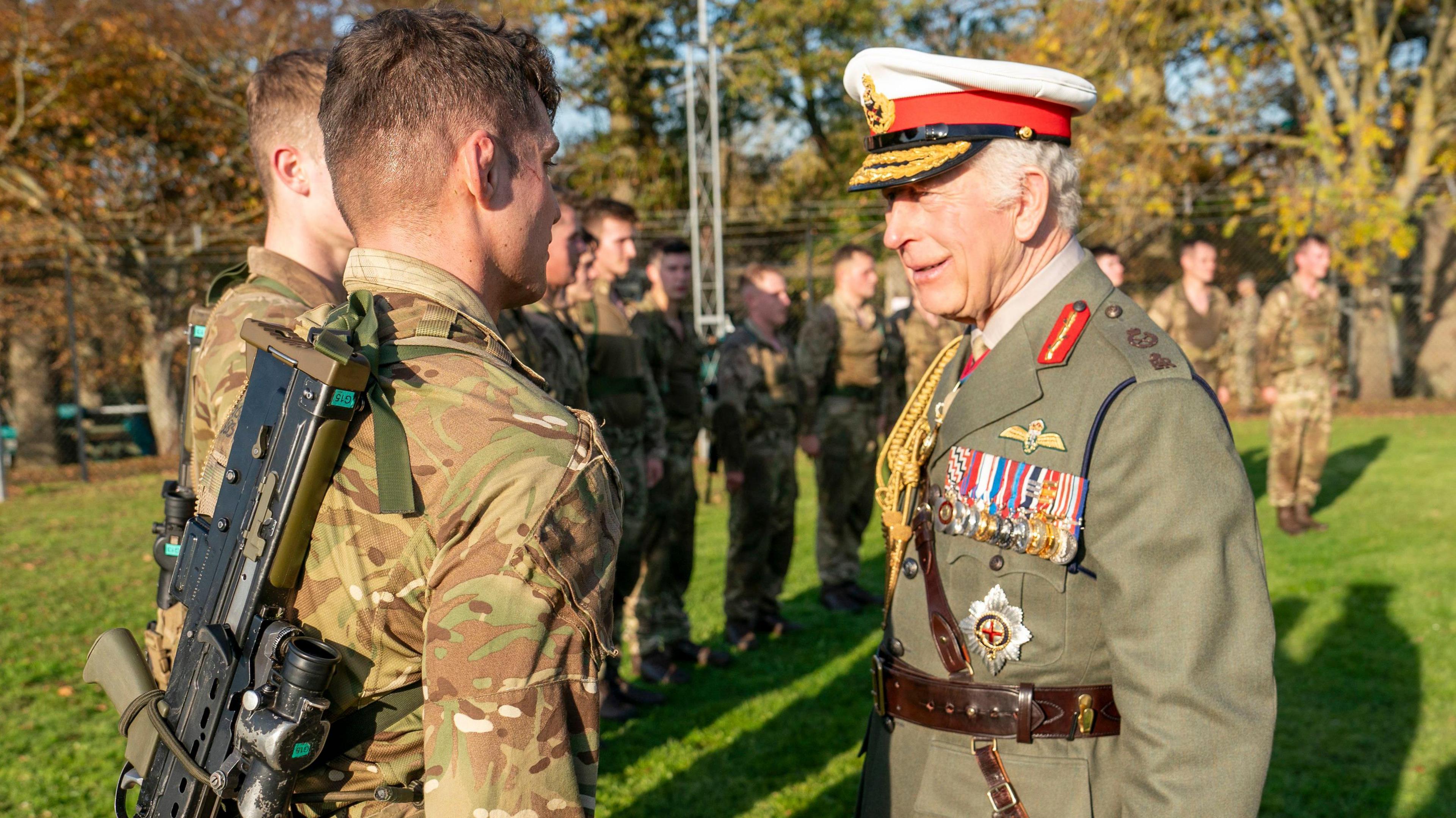 King Charles III wearing military clothing and medals speaking to a line-up of Royal Marine recruits in a field during a royal visit.