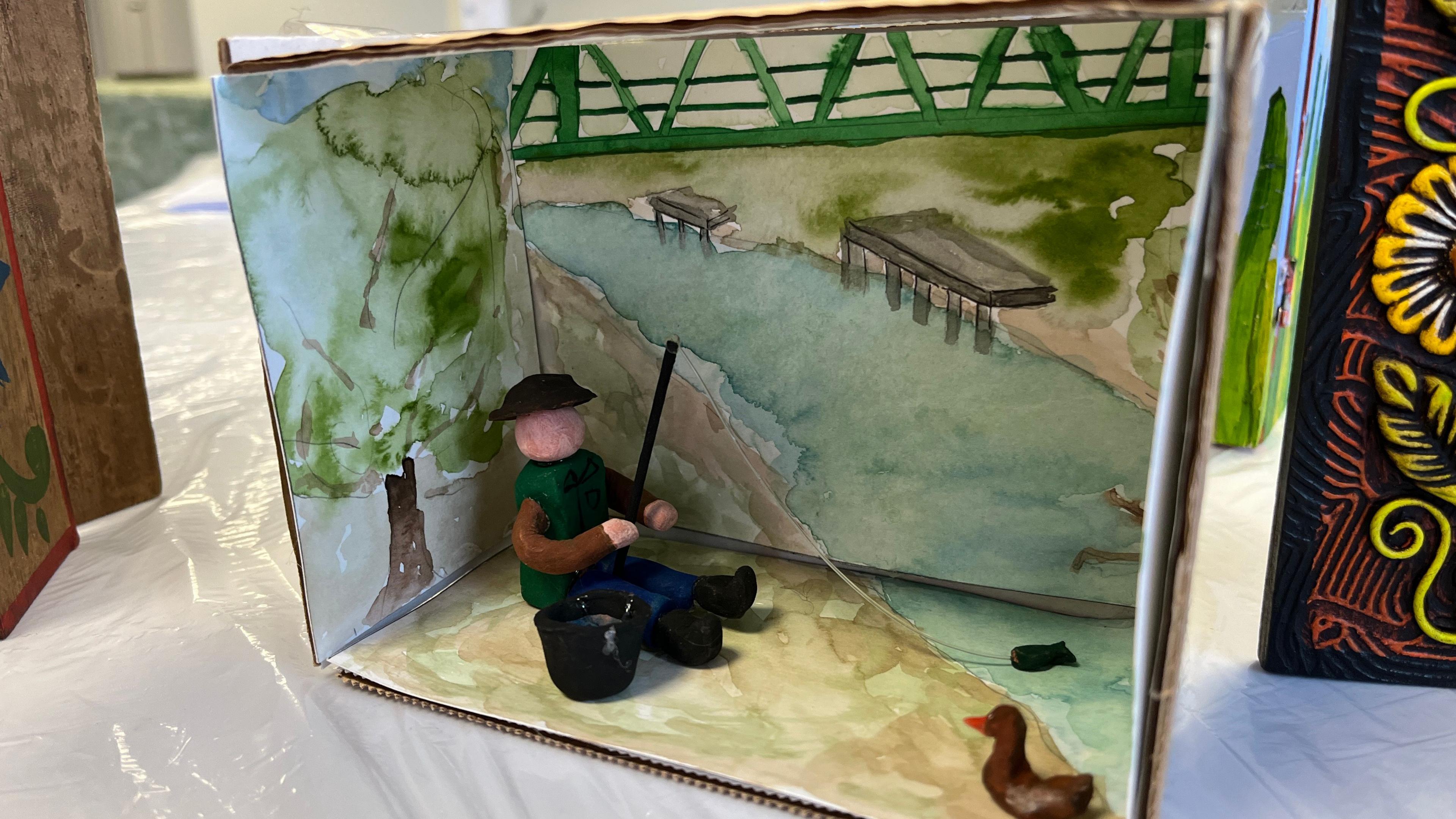 A wooden box has been painted to depict  a nature scene with a river, trees and grass. Two small sculptures are inside the box. One made to look like a person fishing in the painted river and another resembling a duck. A model bucket has also been made as well as a small fish on the end of the fishing rod.