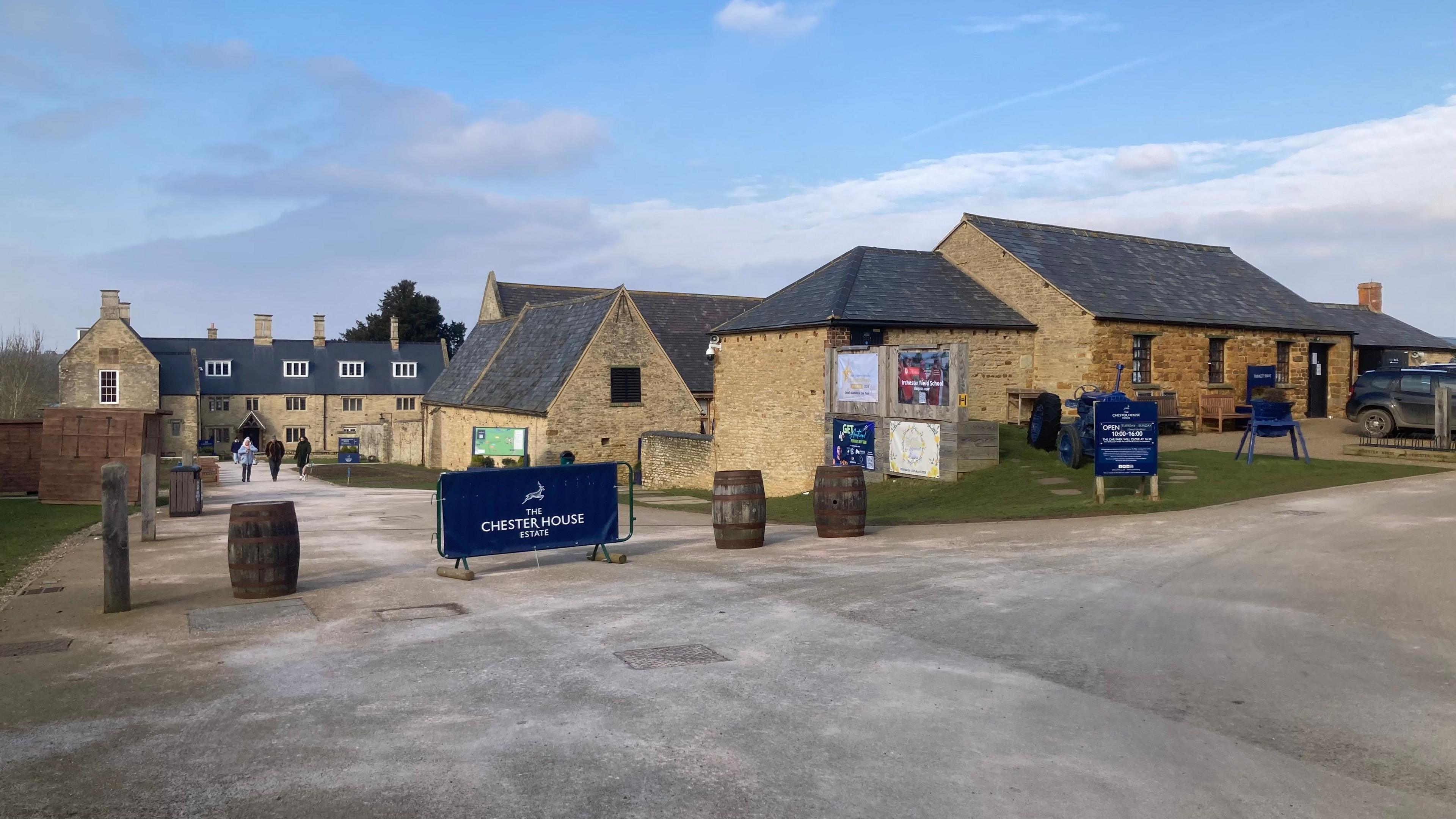 Chester House showing the original three-storey stone farmhouse to the left and other converted buildings housing various exhibits and experiences. There is a CHESTER HOUSE blue banner at the entrance, alongside barrels acting as vehicle barriers.