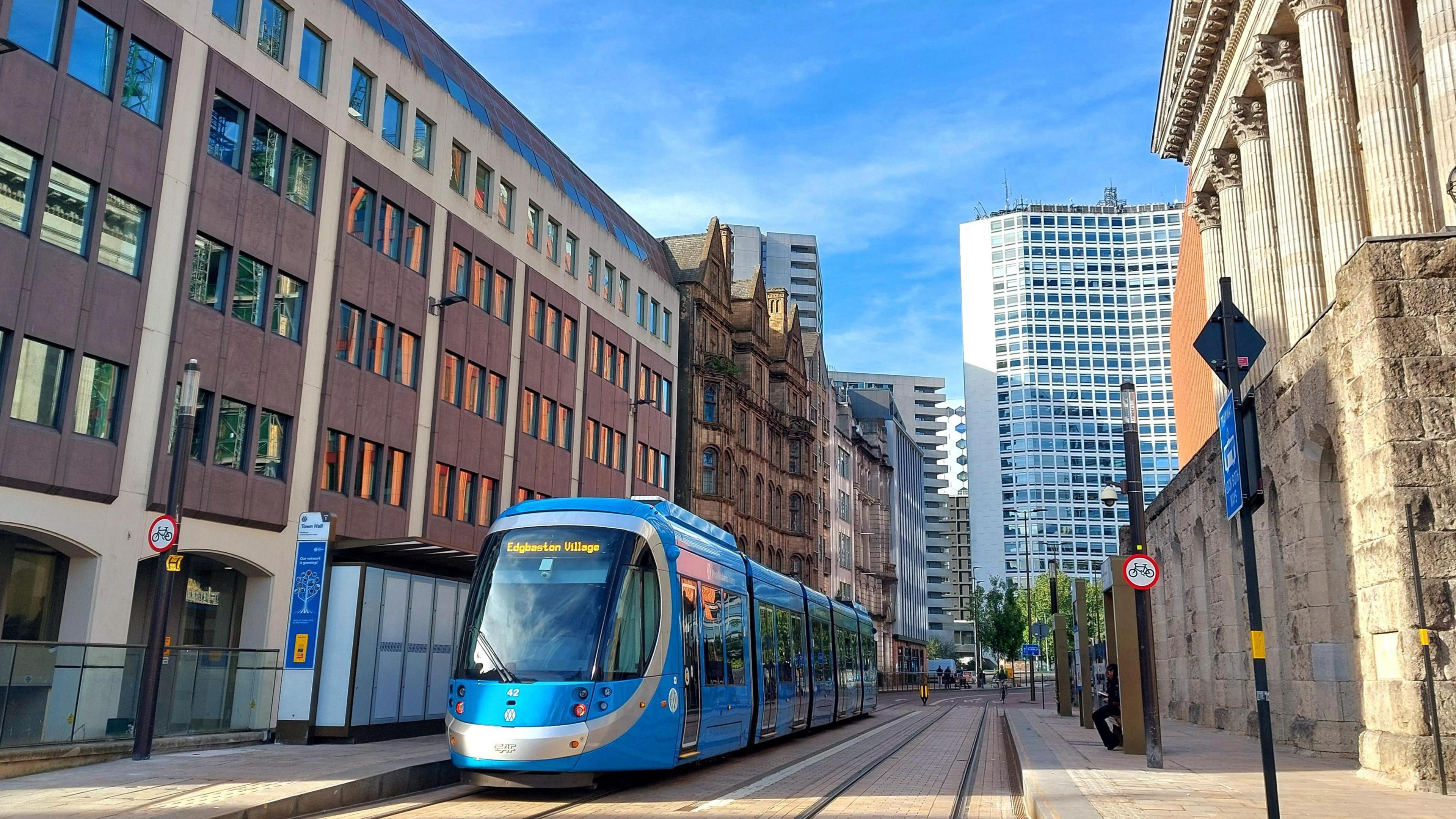 A blue and silver-striped metro tram on its tramlines in Birmingham  between two lines of buildings