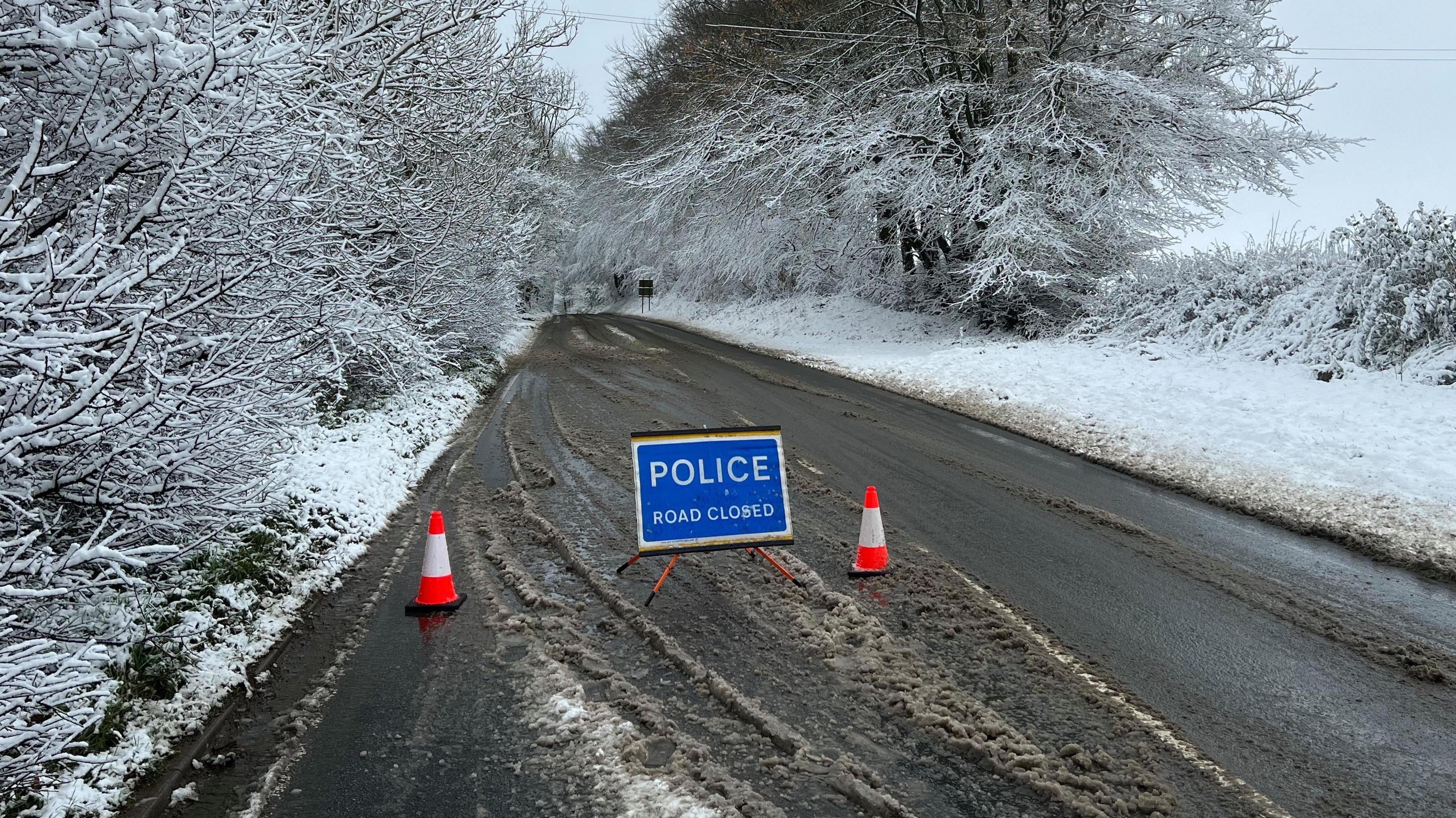 A police 'road closed' sign and two cones blocks a snow and slush covered country road. The hedges and trees either side of the track are covered in a blanket of wet snow.