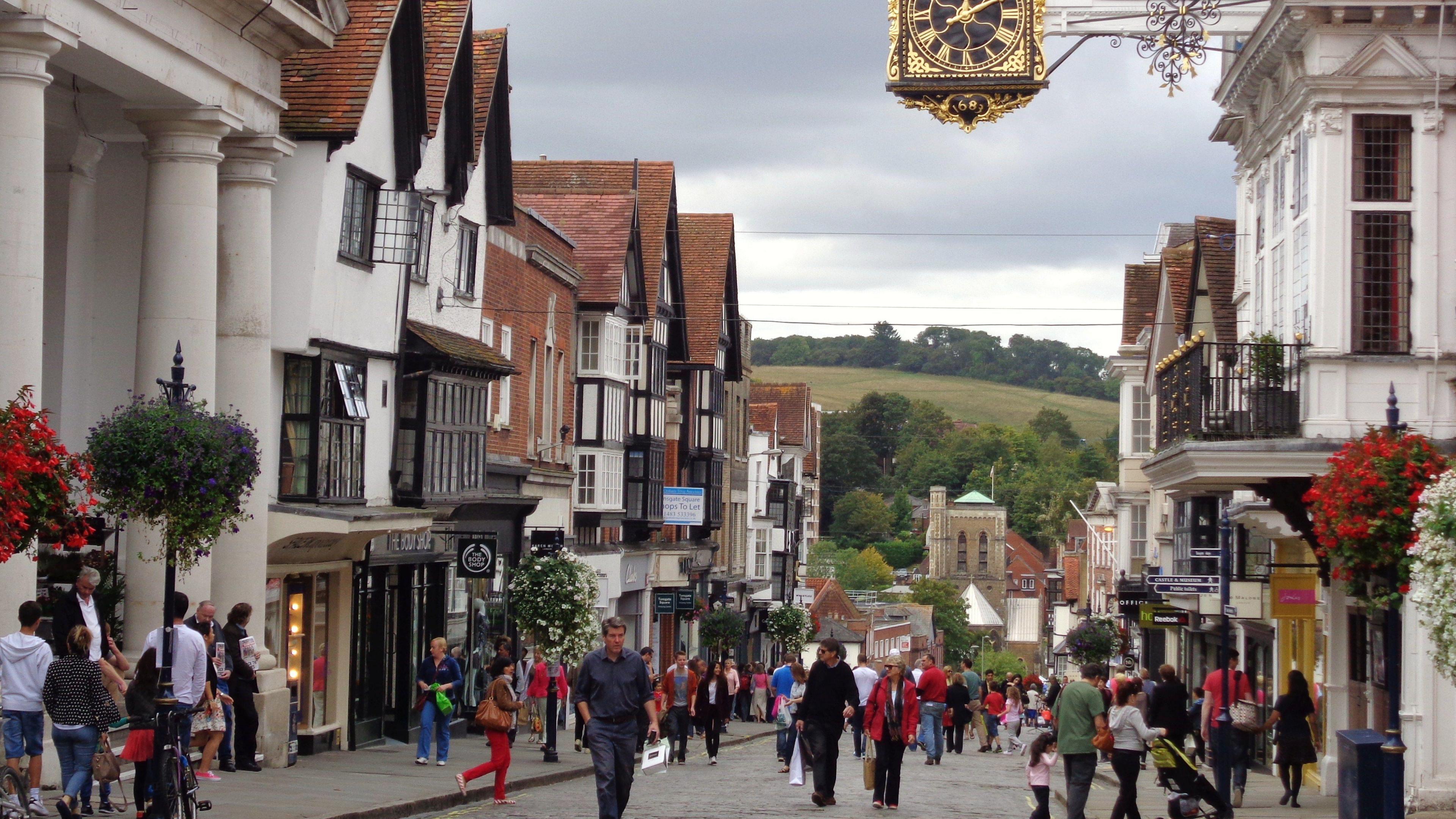 Guildford High Street, close to Tunsgate Arch. There are lots of shoppers on the street. Baskets of flowers are on either side of the road. The Guild Hall clock can be seen at the top of the picture and countryside in the background.