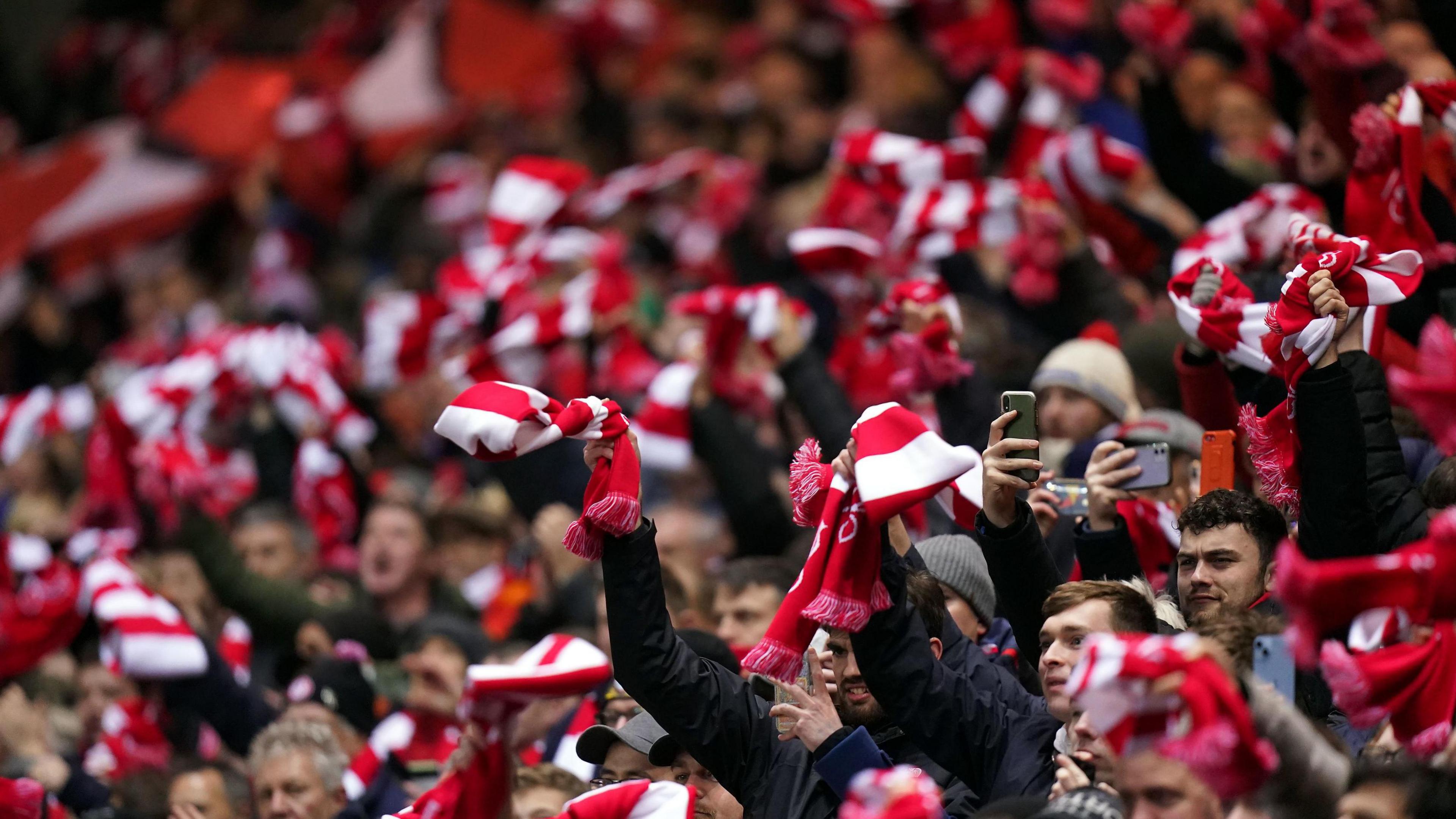 Bristol City fans in the stands at Ashton Gate. Fans wearing and waving red and white scarves. Some in the crowd are holding up phones, indicating they are taking photos or filming.