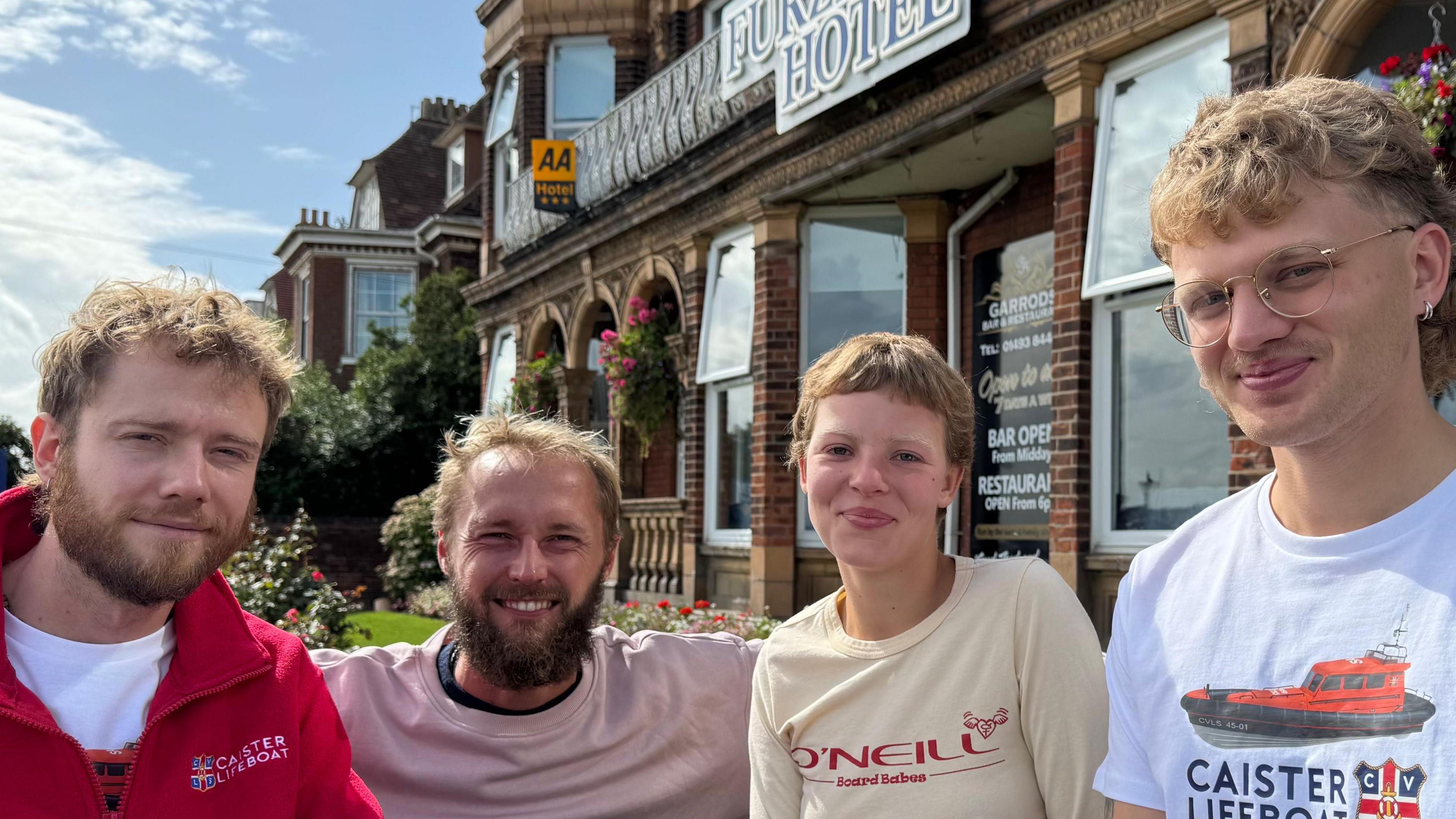 Four people are sitting down next to each other and are looking at the camera smiling. They are sat in front of a building which has its windows open and a sign  with writing on. 