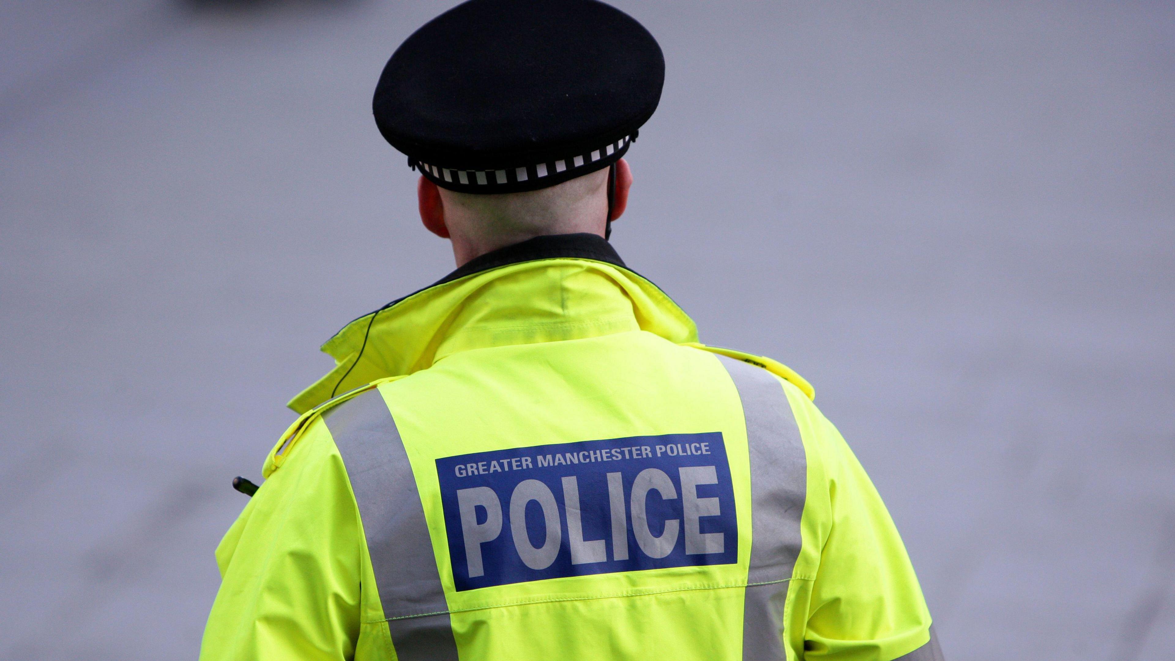 A police officer from Greater Manchester Police on duty in the city centre wearing a green high-vis jacket emblazoned with the force's name and wearing a place hat, facing away from the camera.