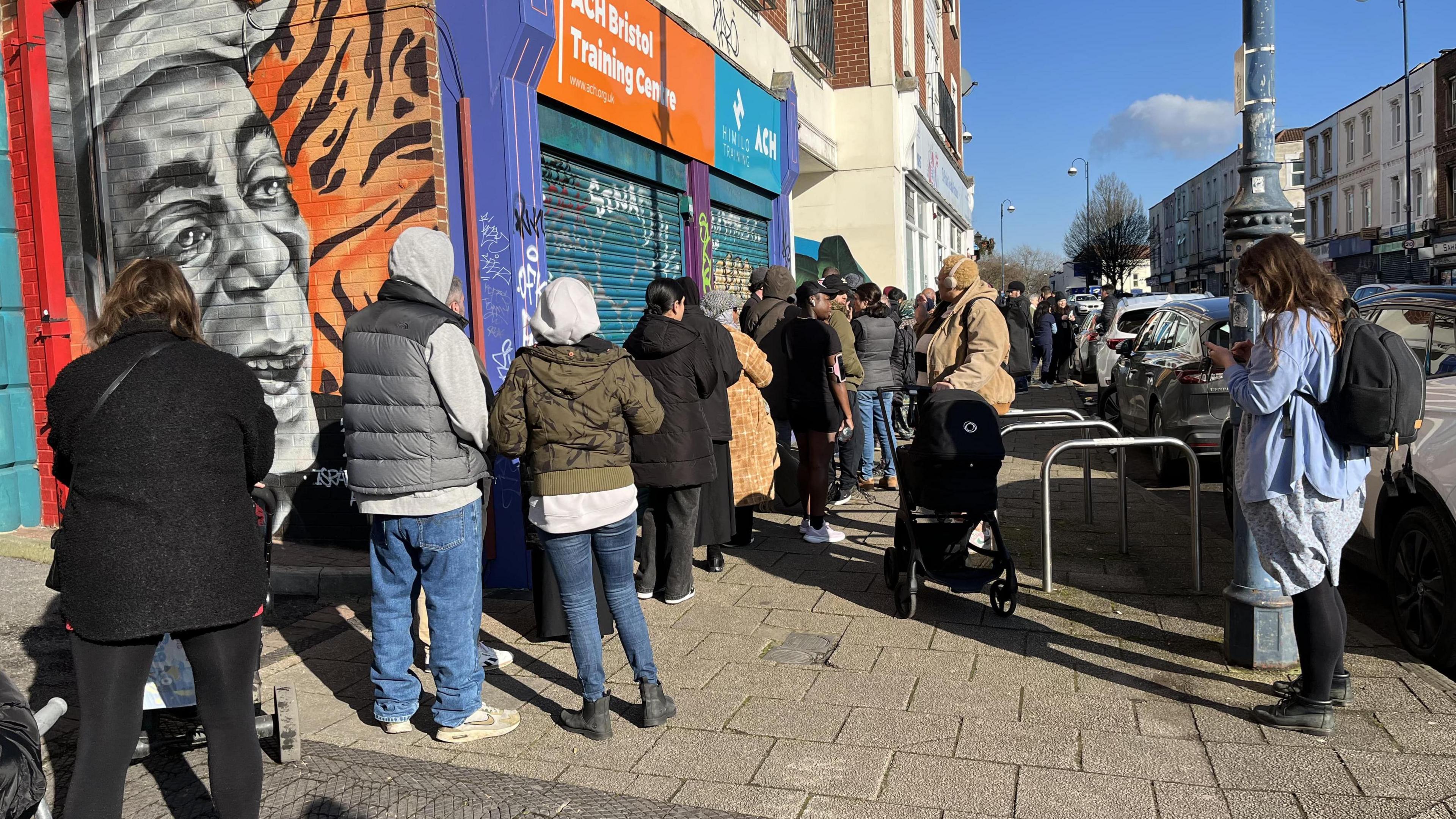 A queue of people standing outside St Paul's Dental Surgery in Bristol. The surgery is on a high street and the queues are going down the street. There is around 30 to 40 people waiting in the queue.