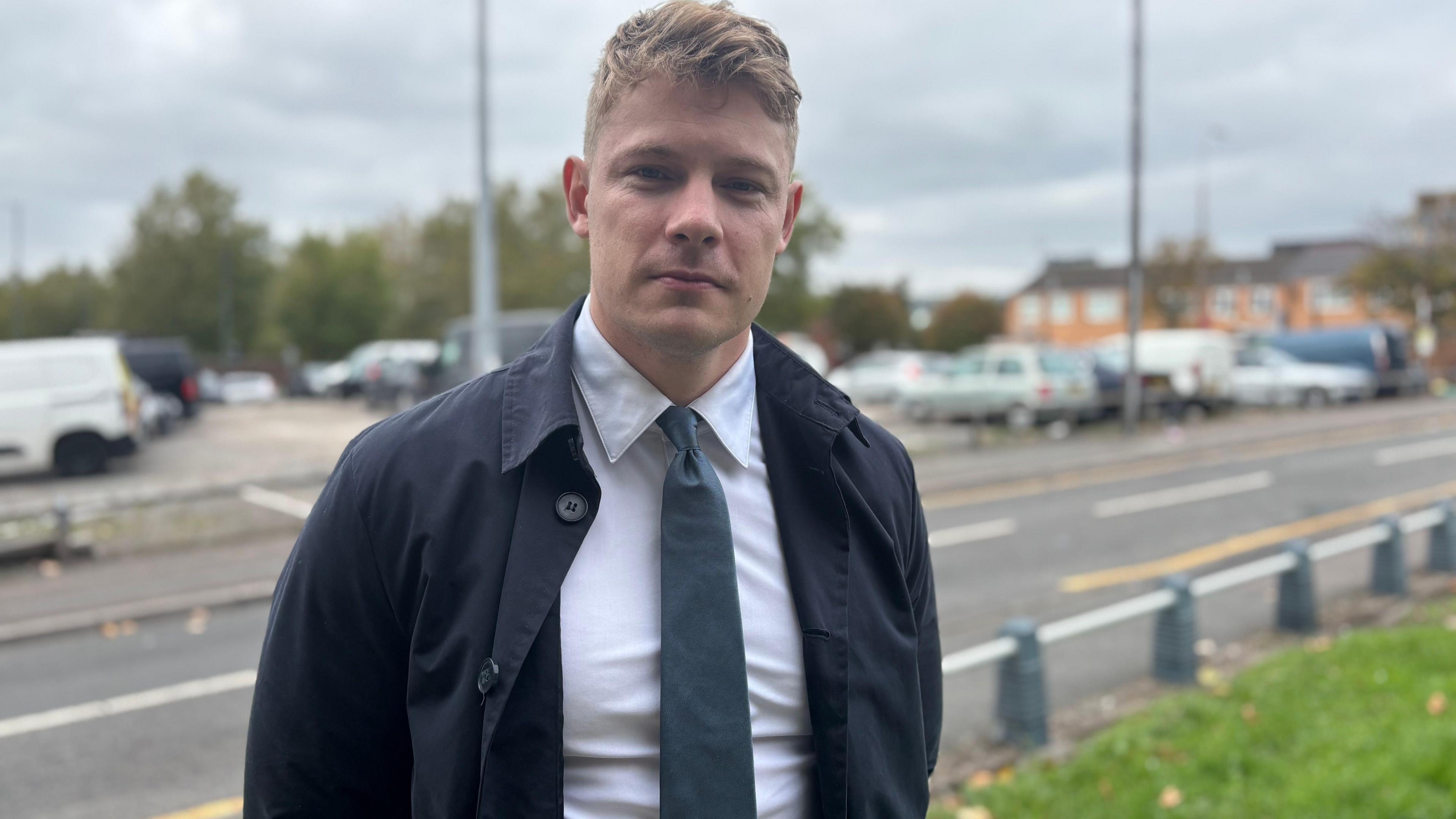 Carl Cashman, who has blond hair and wears a navy blue jacket, white shirt and a blue/grey tie, smiles for the camera as he is photographed in front of a residential road.