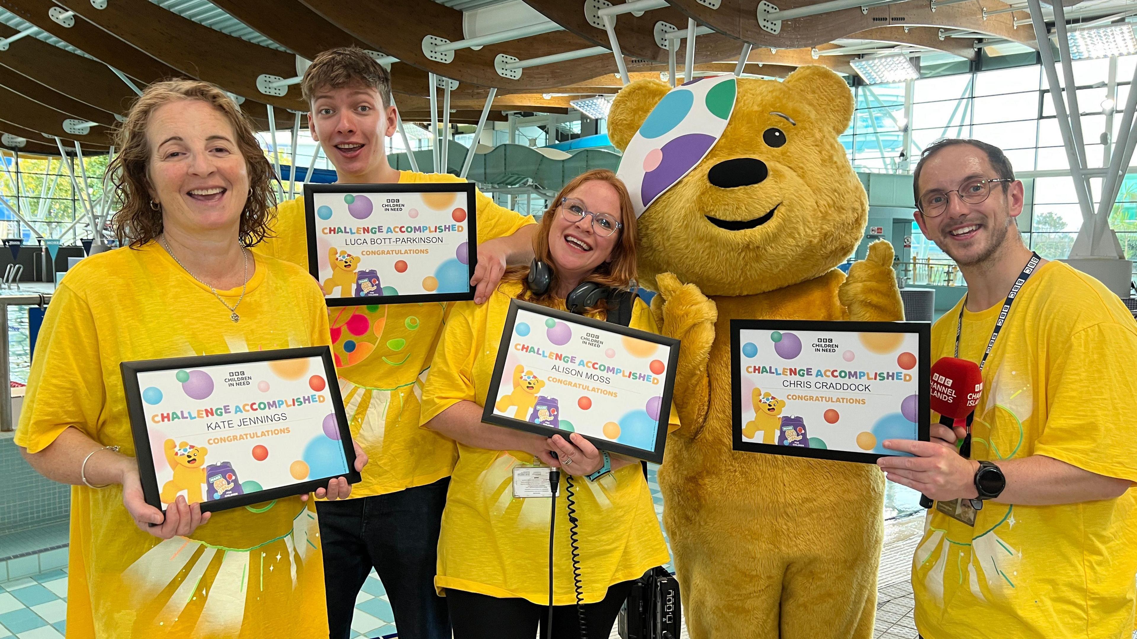 The team hold framed certificates and smile at the camera. They're all wearing top tops with Pudsey's face on them. Pudsey is standing with them giving a thumbs up. Behind them is the pool and there is a big slide in the background.