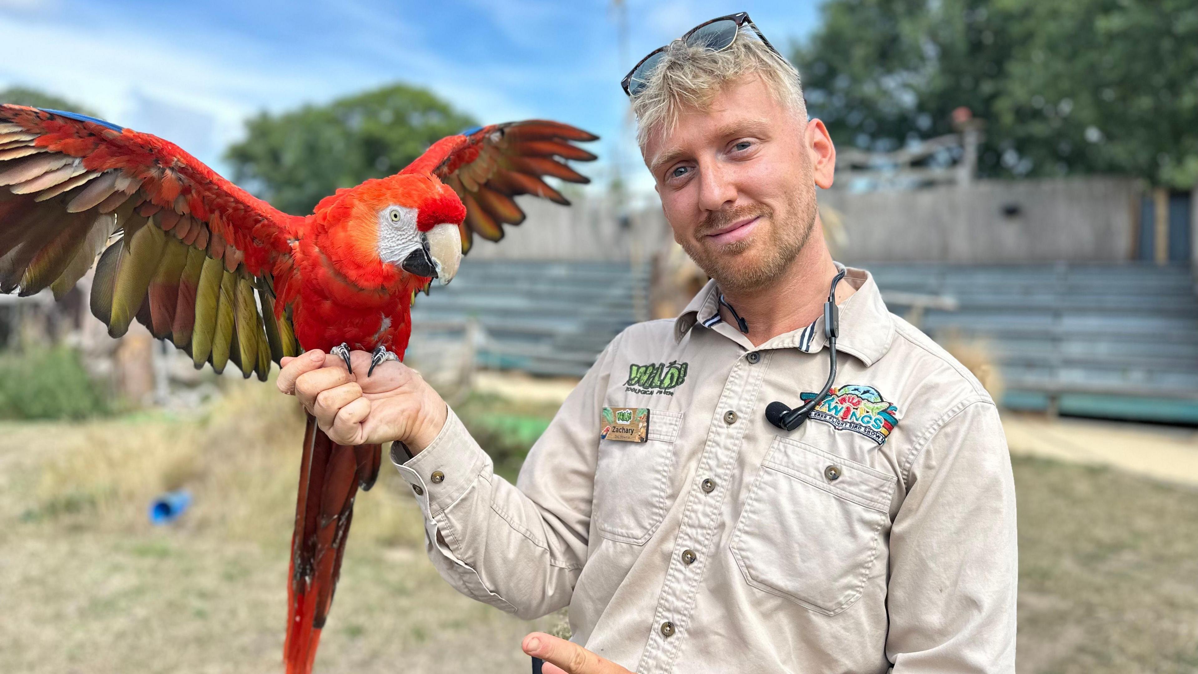 Zachary Hollinshead is wearing a safari material top and has short blonde/brown hair and sunglasses on his head.