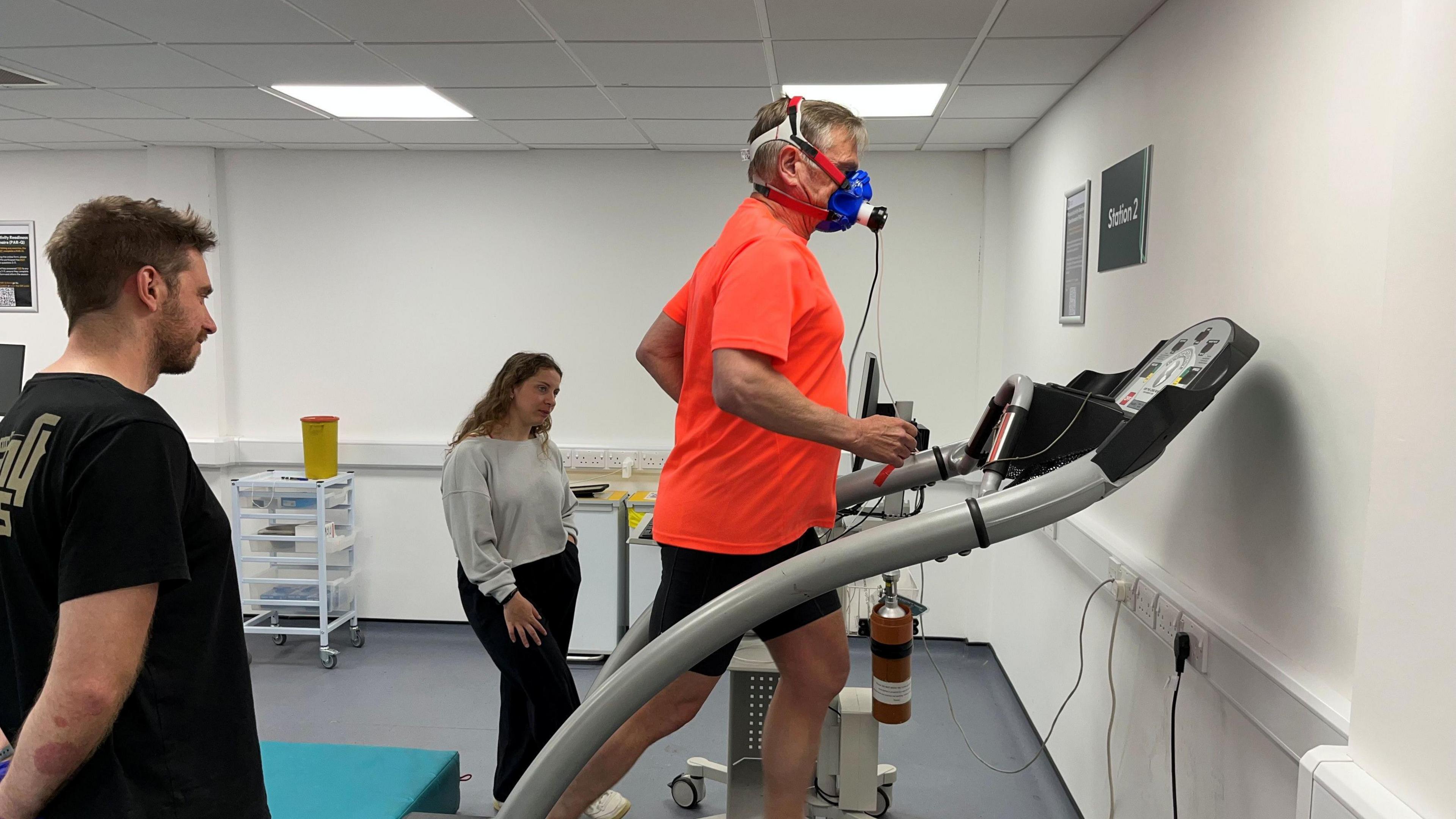A man running on a machine wearing an orange shirt and a mask. He is surrounded by two people monitoring him.