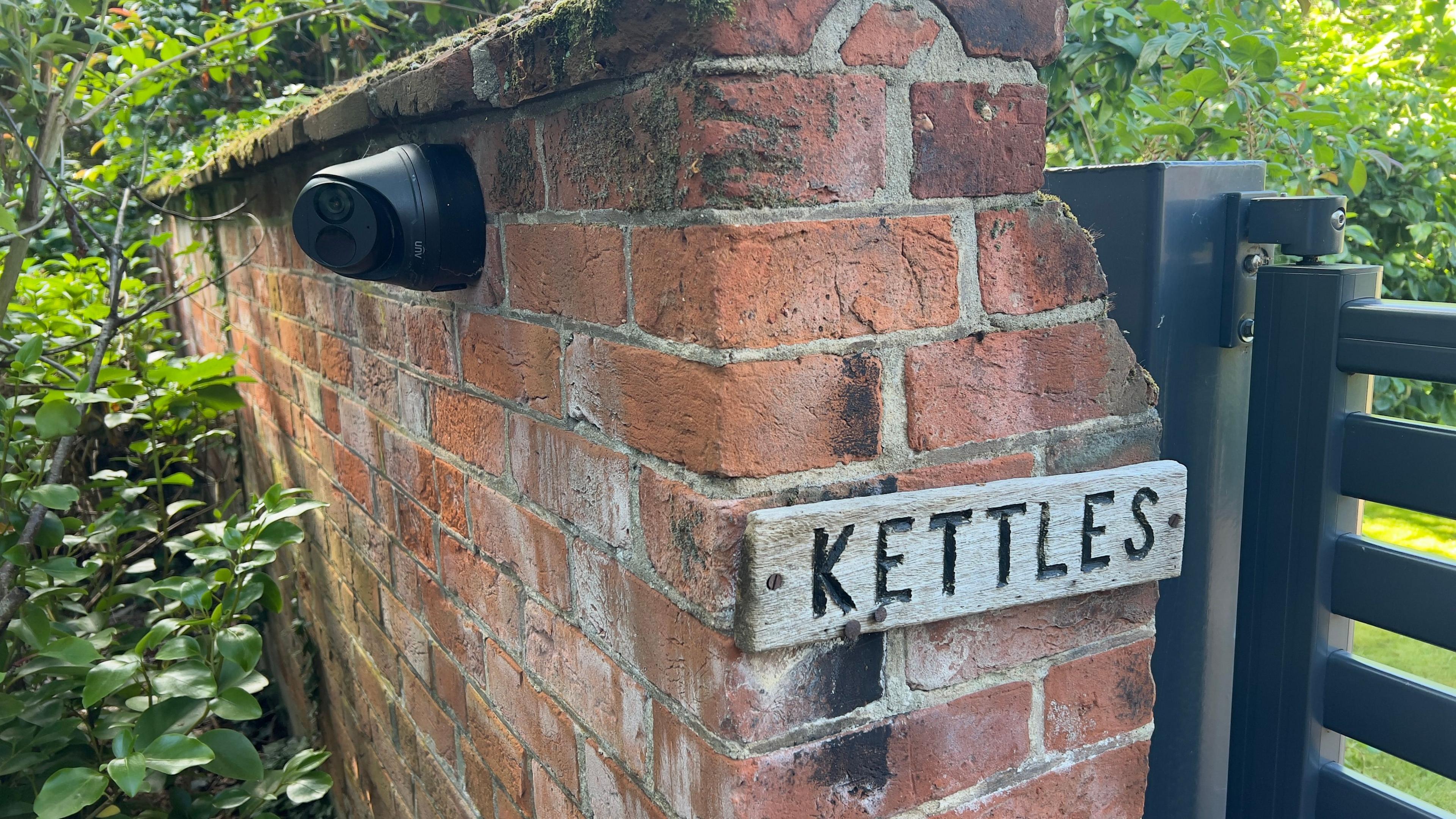 A brick wall with the sign 'Kettles' next to a brown wooden gate. On the other side is a big green bush