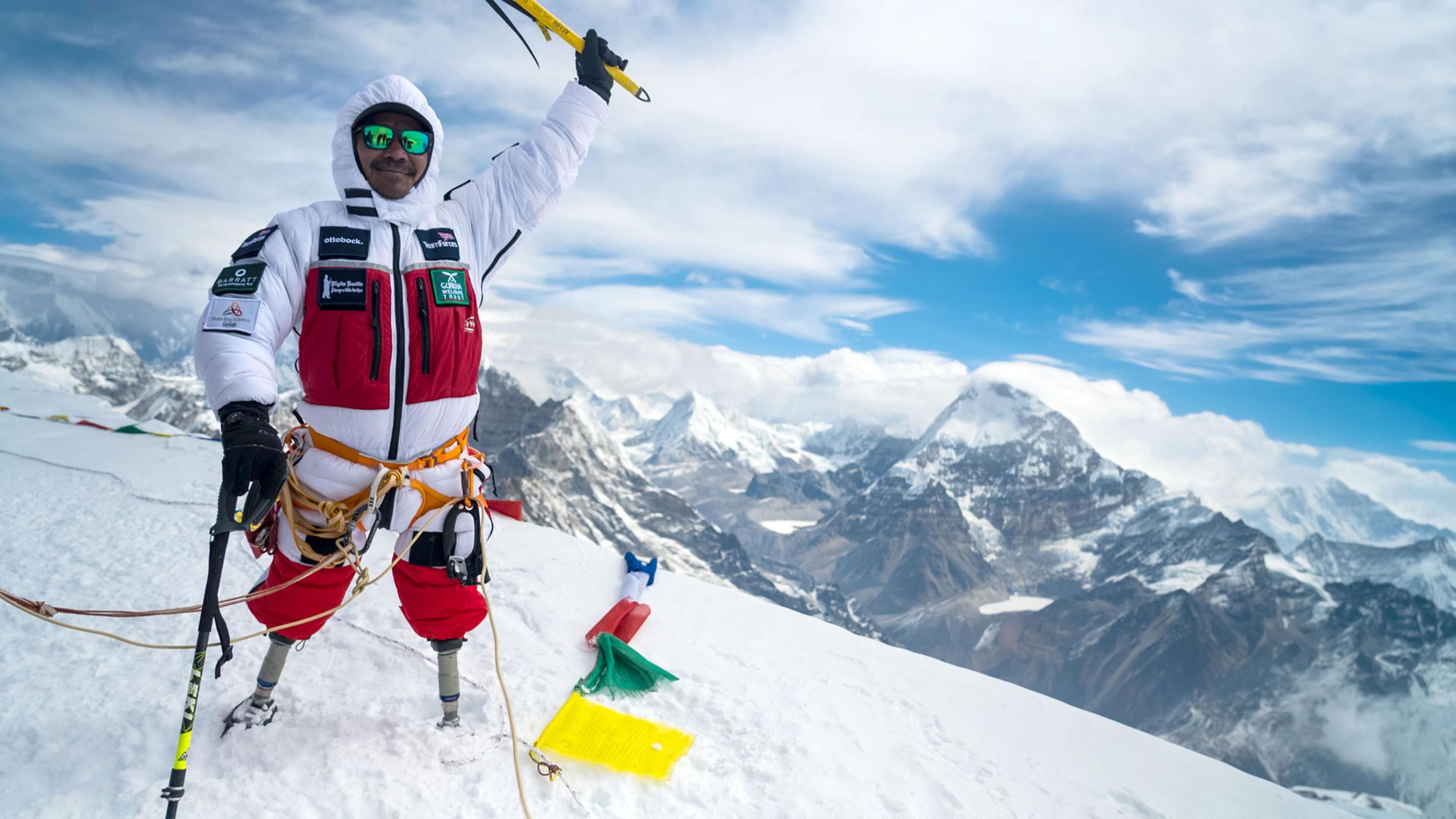 A man in a red and white snow suit, with two above knee prostheses stands atop Mount Everest holding his hand in the air to celebrate. 