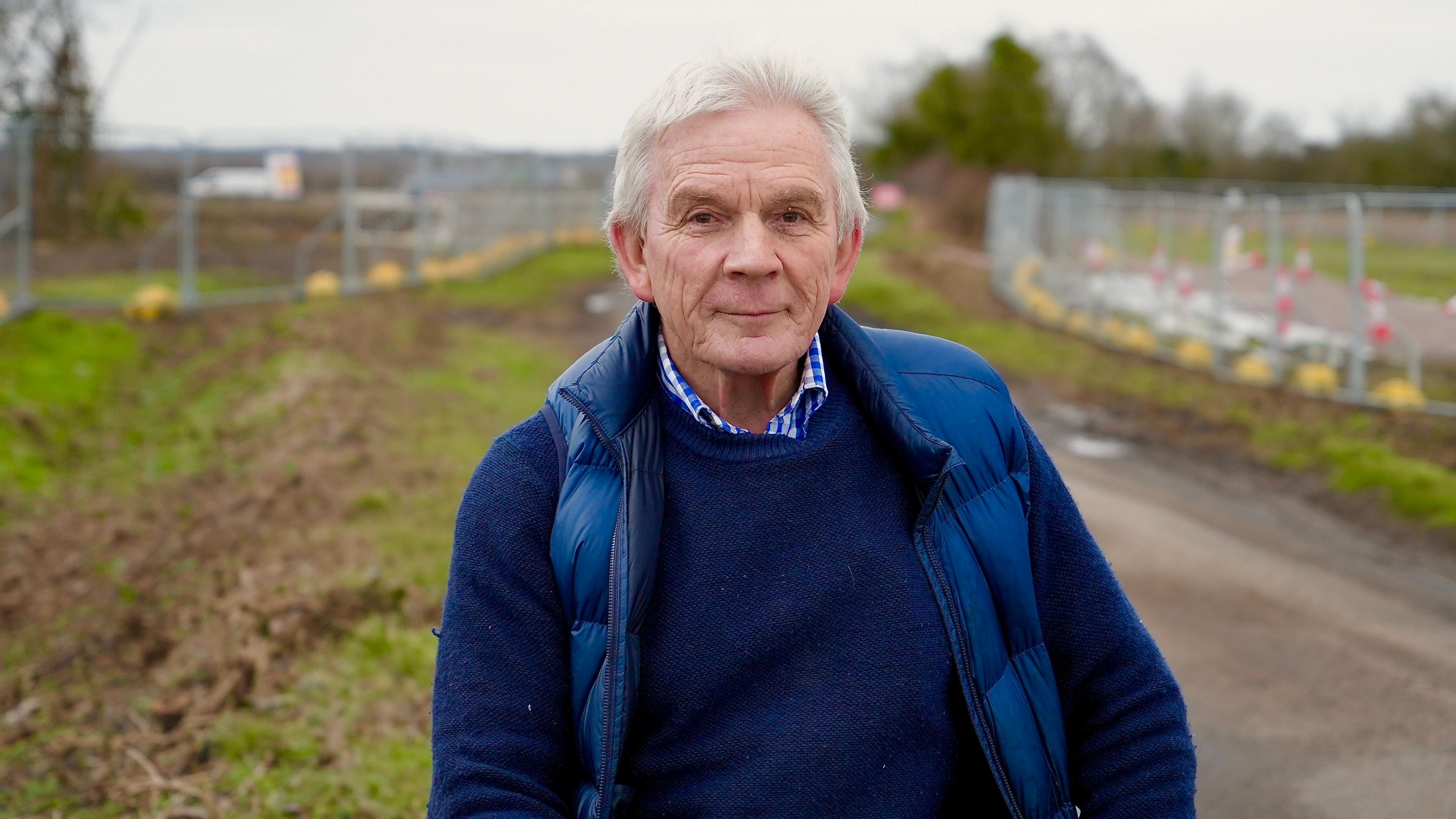 David Grant standing next to fields compulsorily purchased by Sizewell C. He has white hair and is wearing a blue gilet over a blue sweater and blue and white checked shirt. Fences and cones can be seen behind him.