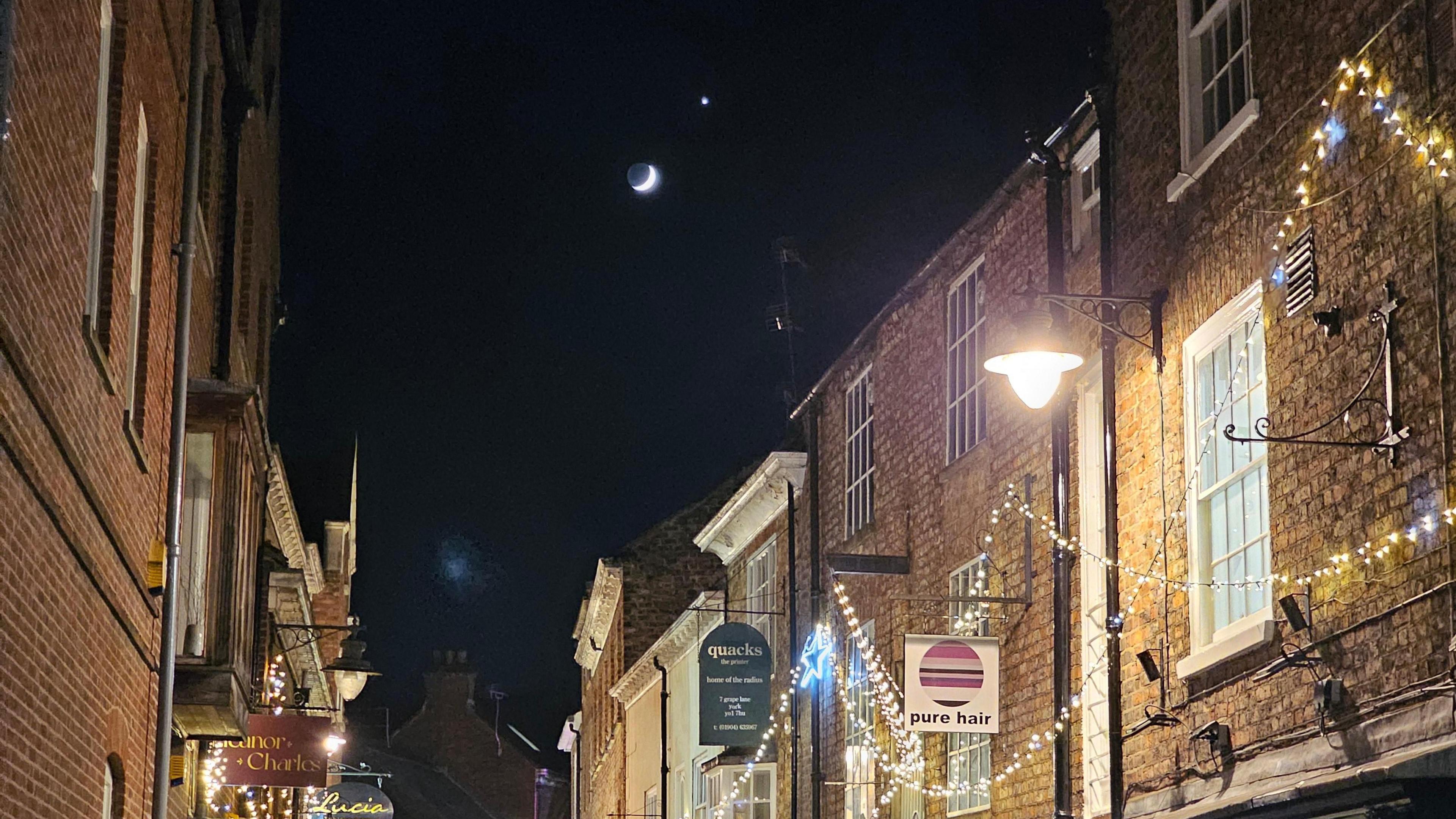 The moon and Venus shine down on a street in York. Stings of fairy lights are threaded alongthe buildings. Signs for Pure Hair and Quacks shop are visible. 