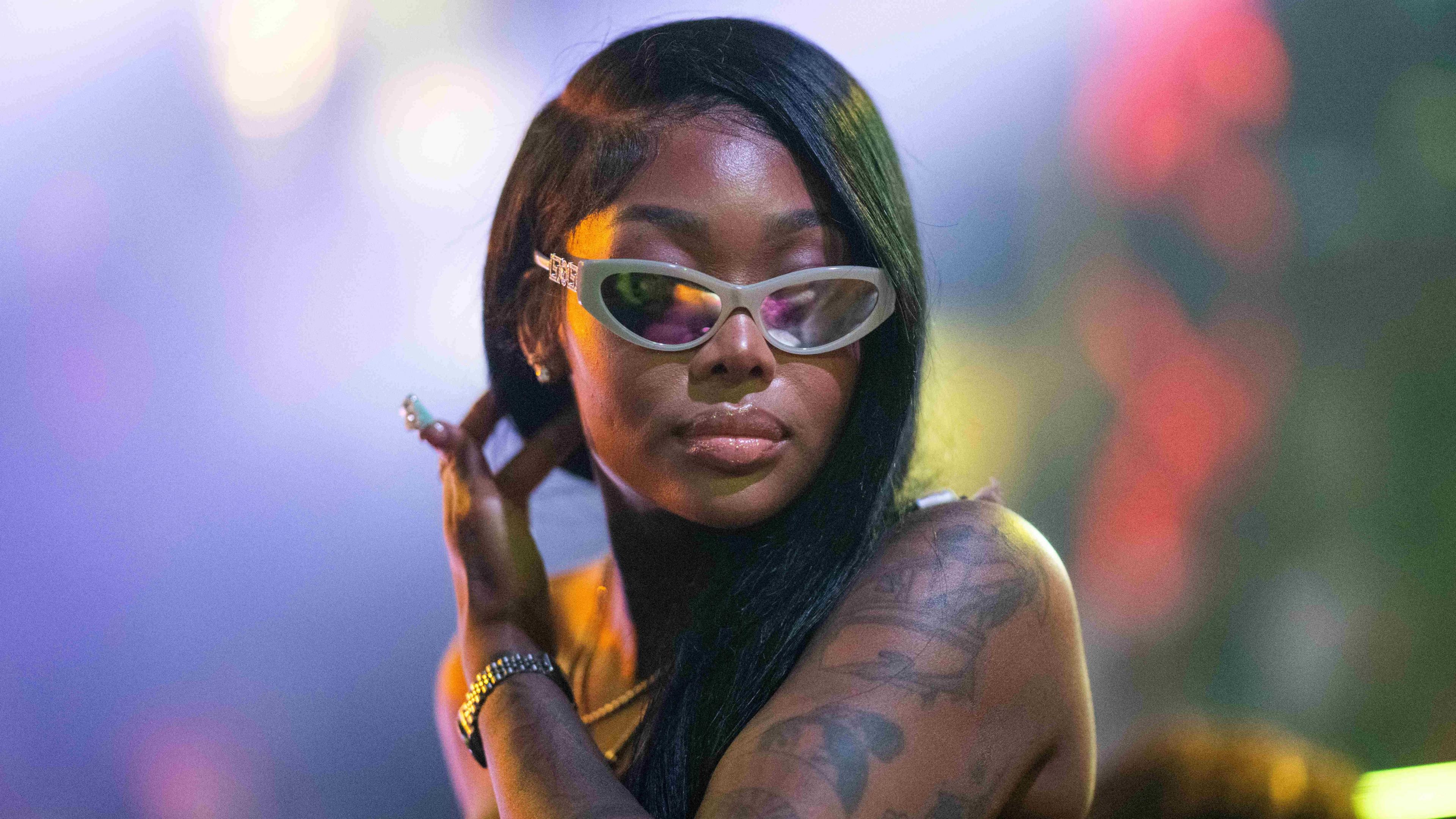 Summer Walker wears a one-armed black top, revealing a heavily tattooed left arm. She has bobbed black hair and wears reflective sunglasses, her left hand running through her hair.
