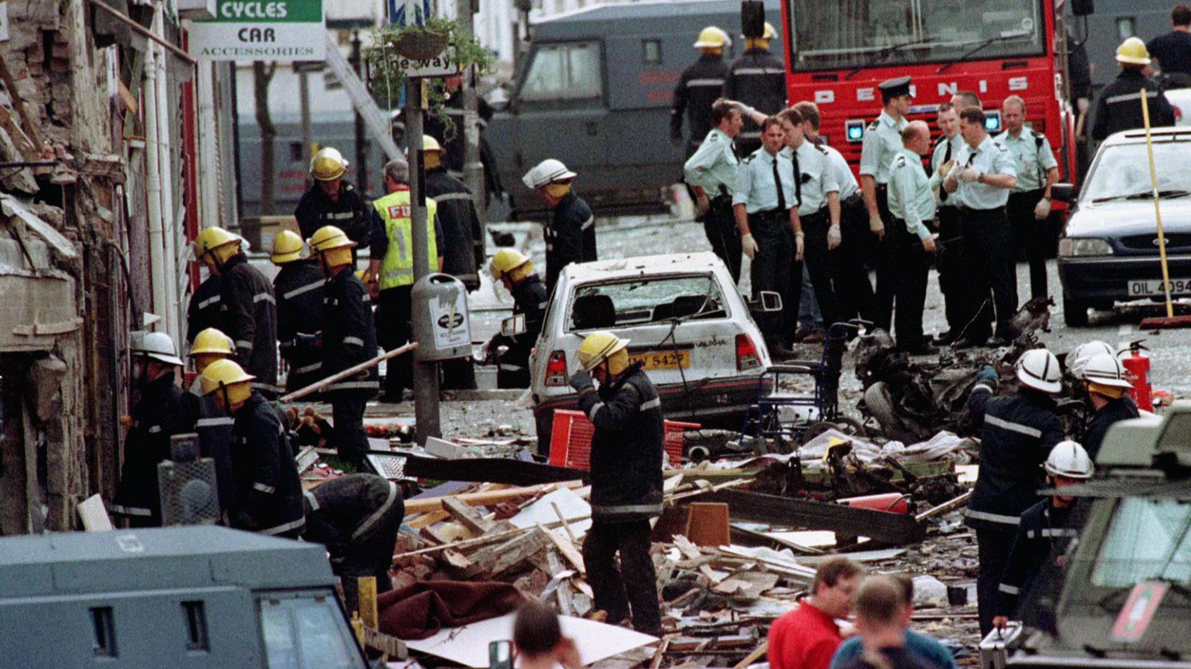 Several emergency responders are standing in a street in the aftermath of a bomb. Rubble and debris cover the street and road. Cars are damaged with glass blown out