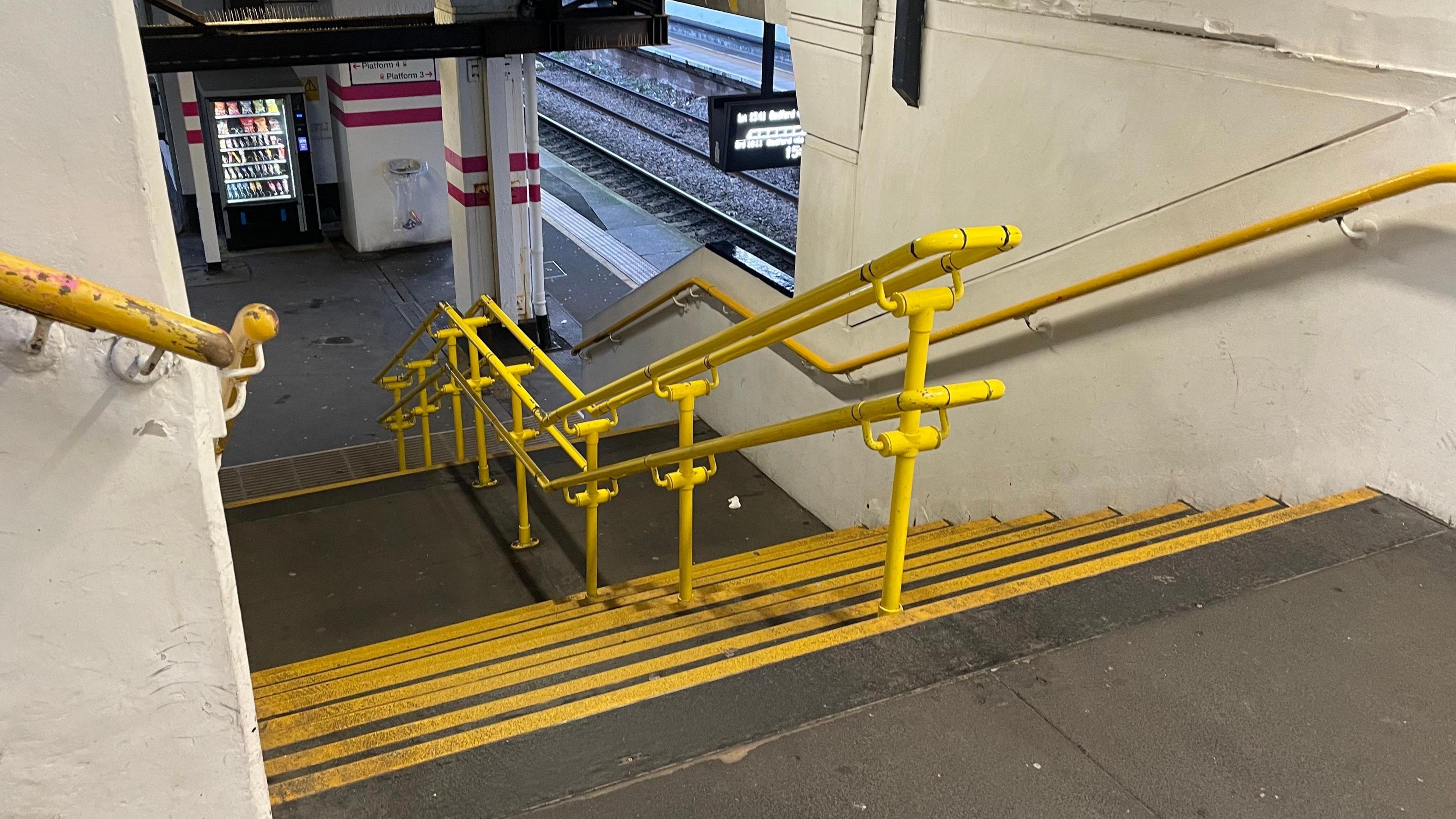 Looking down an empty flight of stairs at Luton station. There are yellow, metal handrails along the walls and a central yellow handrail running along the middle of the flight. Each step has a yellow kick guard on its edge. At the bottom of the stairs a vending machine can be seen on the platform as well as a glimpse of the railway tracks and an information board at the bottom.