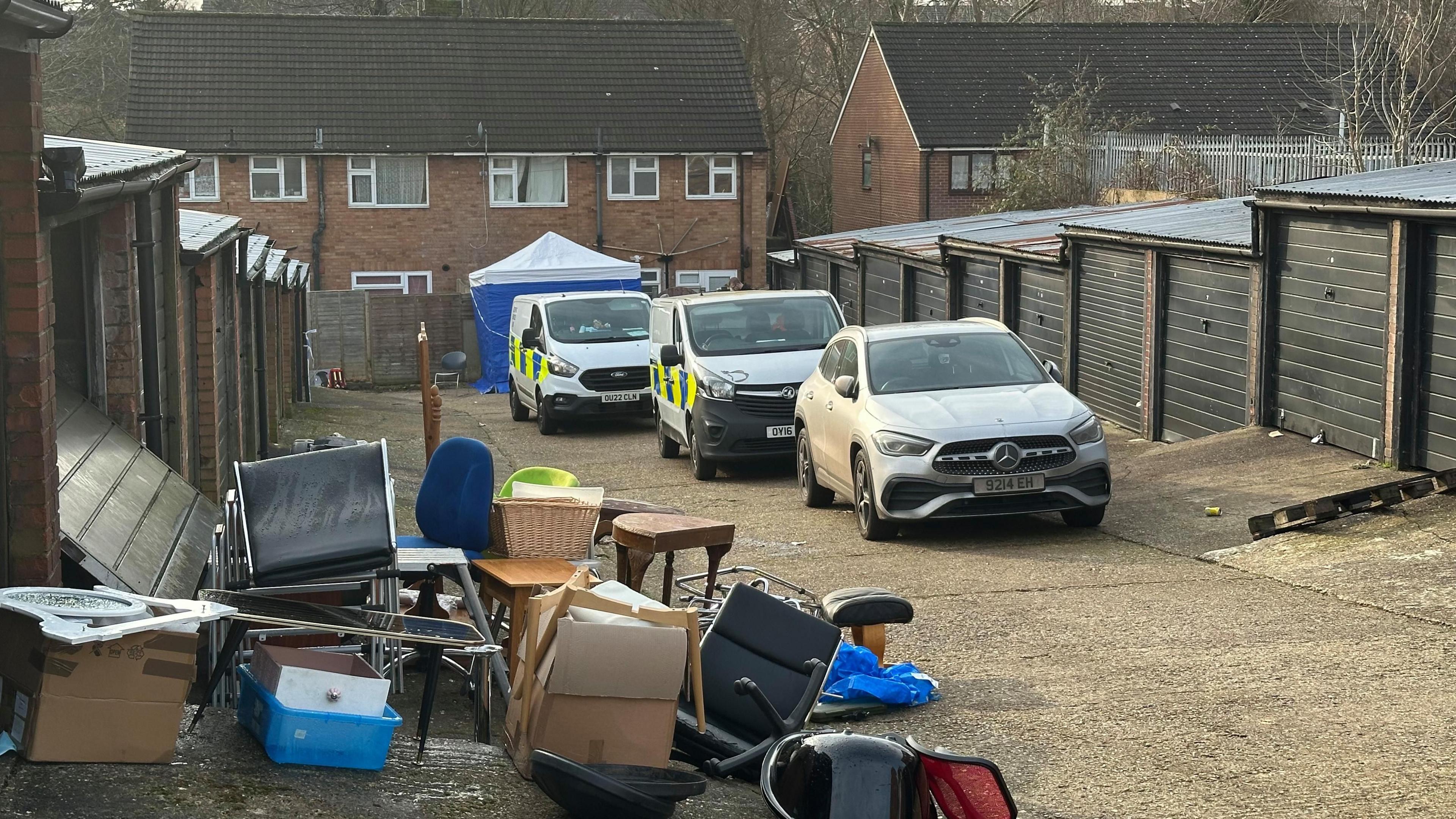 A set of garages behind a block of flats or houses, with two police vans parked at one end. In front of one garage is a pile of furniture and boxes.