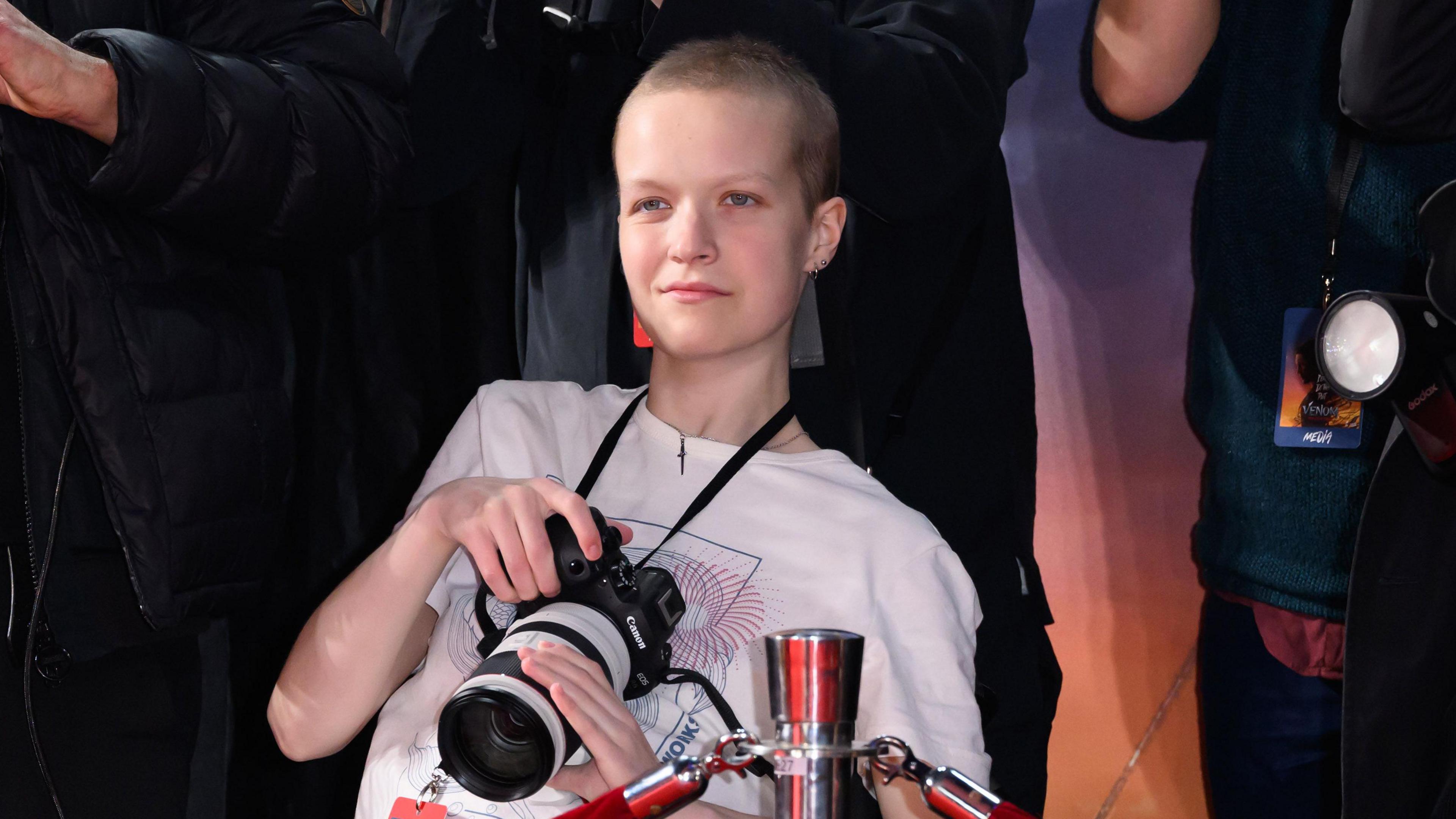 A teenage girl with very short brown hair wearing a white T shirt holding a professional Canon camera surrounded by press photographers