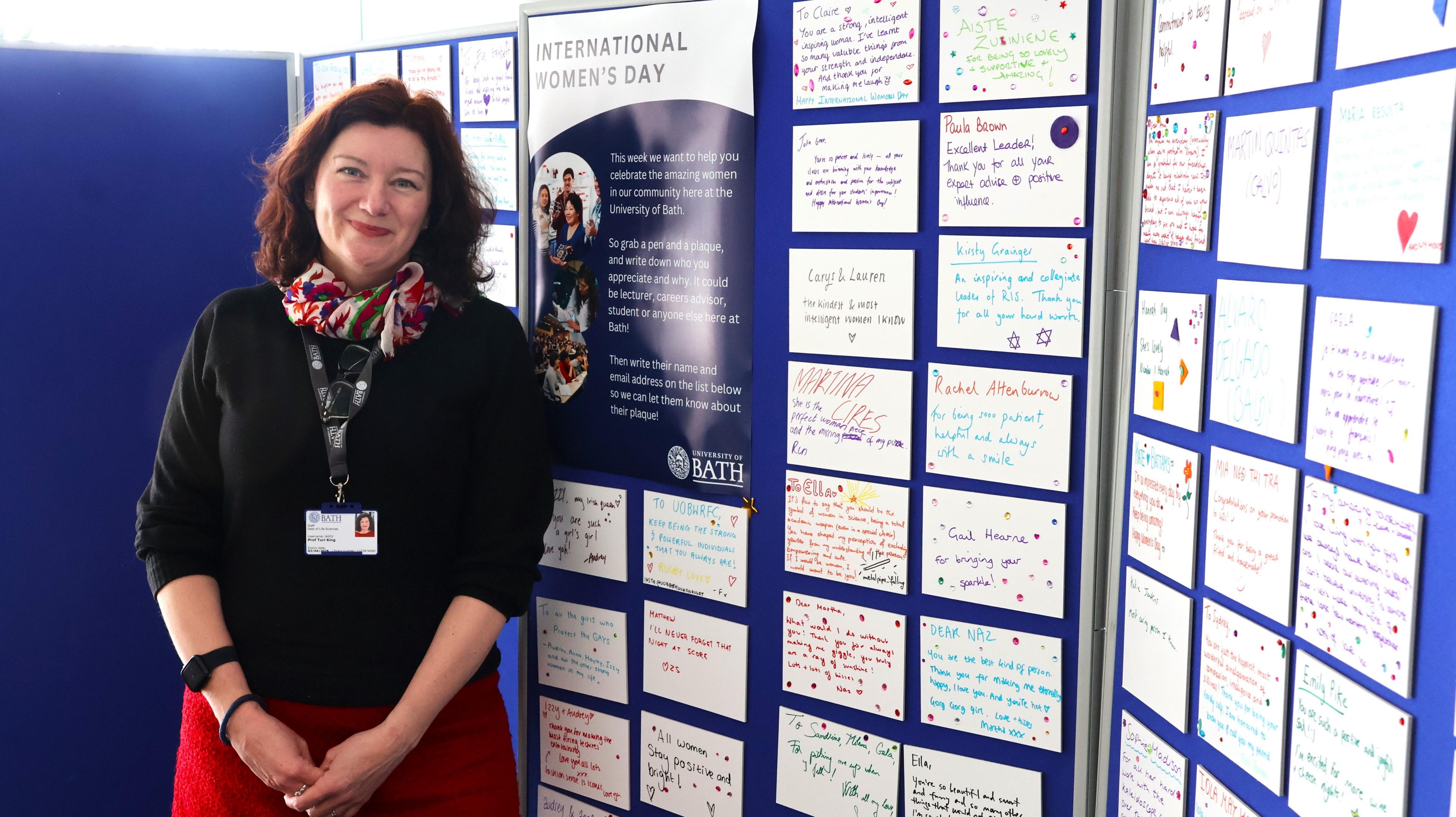 Prof Turi King stands in front of the wall of plaques dedicated to the women at the university. She has short reddish-brown hair, a black top, a red skirt and is smiling at the camera.