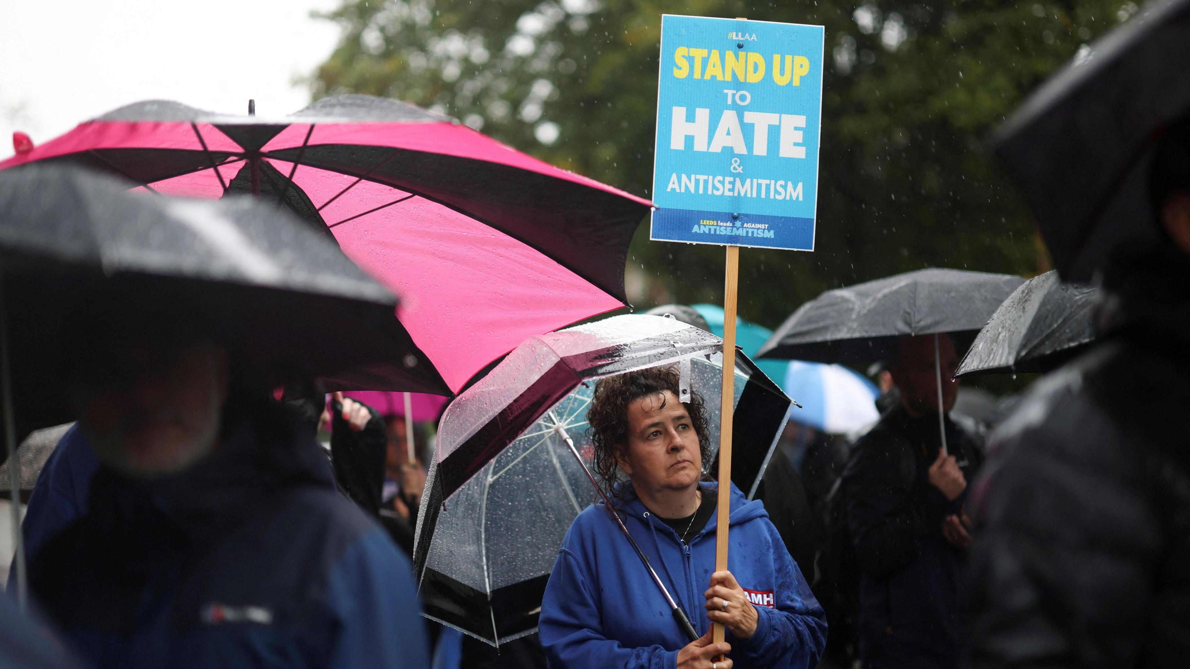 A woman in a blue top holds a blue placard saying Stand Up To Hate And Antisemitism. She is surrounded by other people at a vigil where they hold umbrellas in the rain.