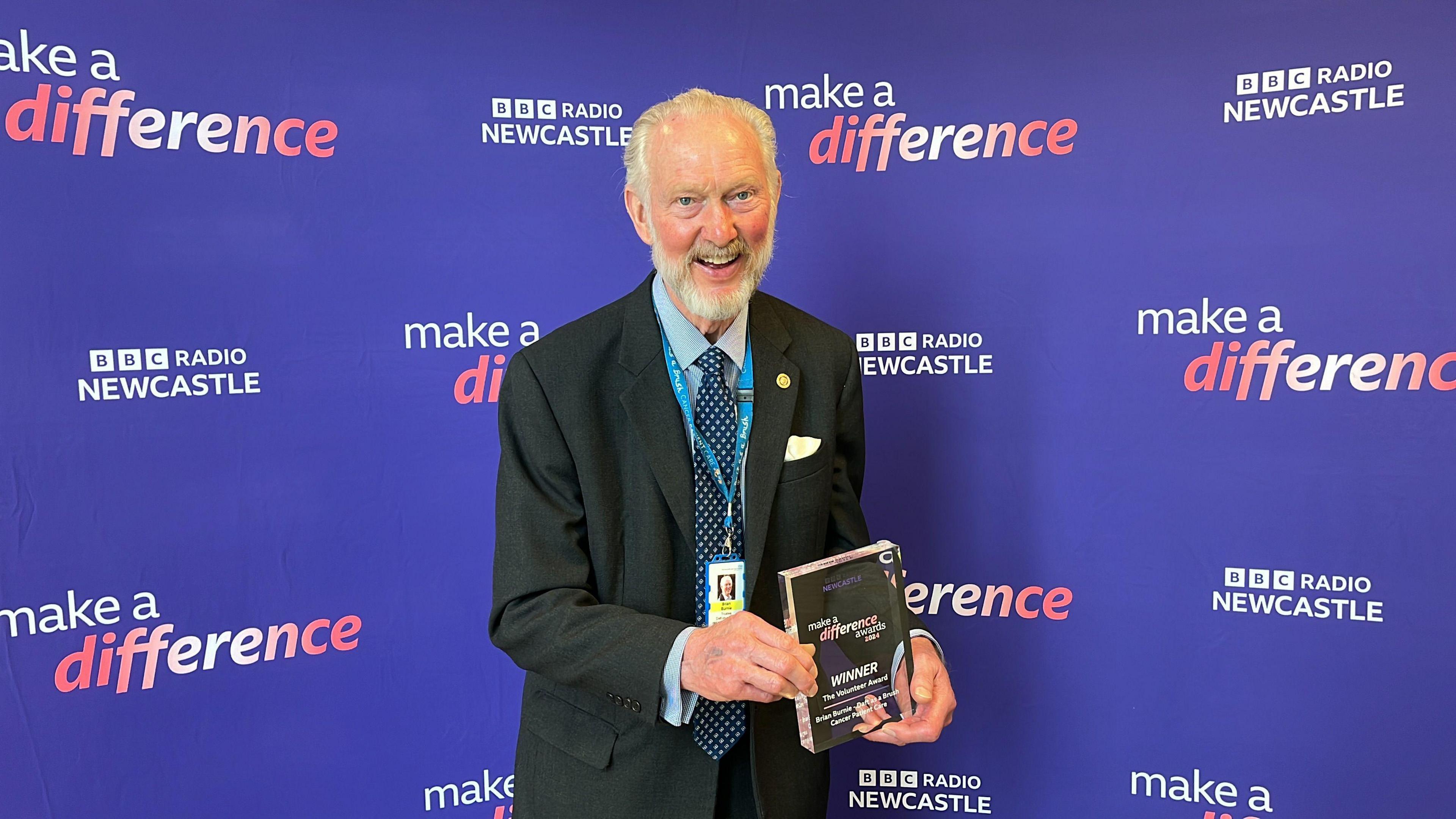 Brian Burnie smiling and holding his award. He has grey hair and a beard.