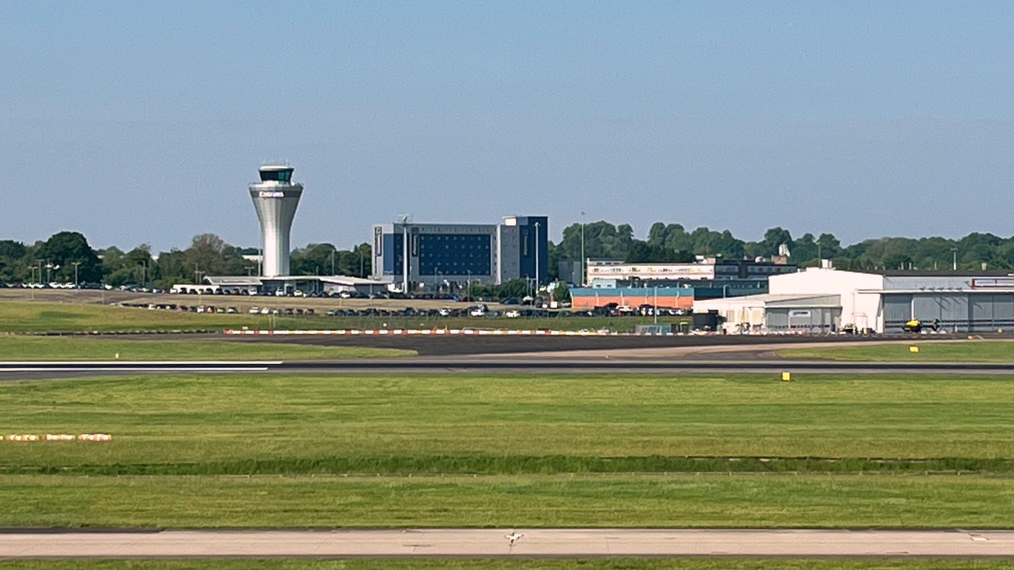 A runway at Birmingham Airport, surrounded by grass. Airport buildings, including a control tower, are seen in the distance in front of green trees on the horizon.