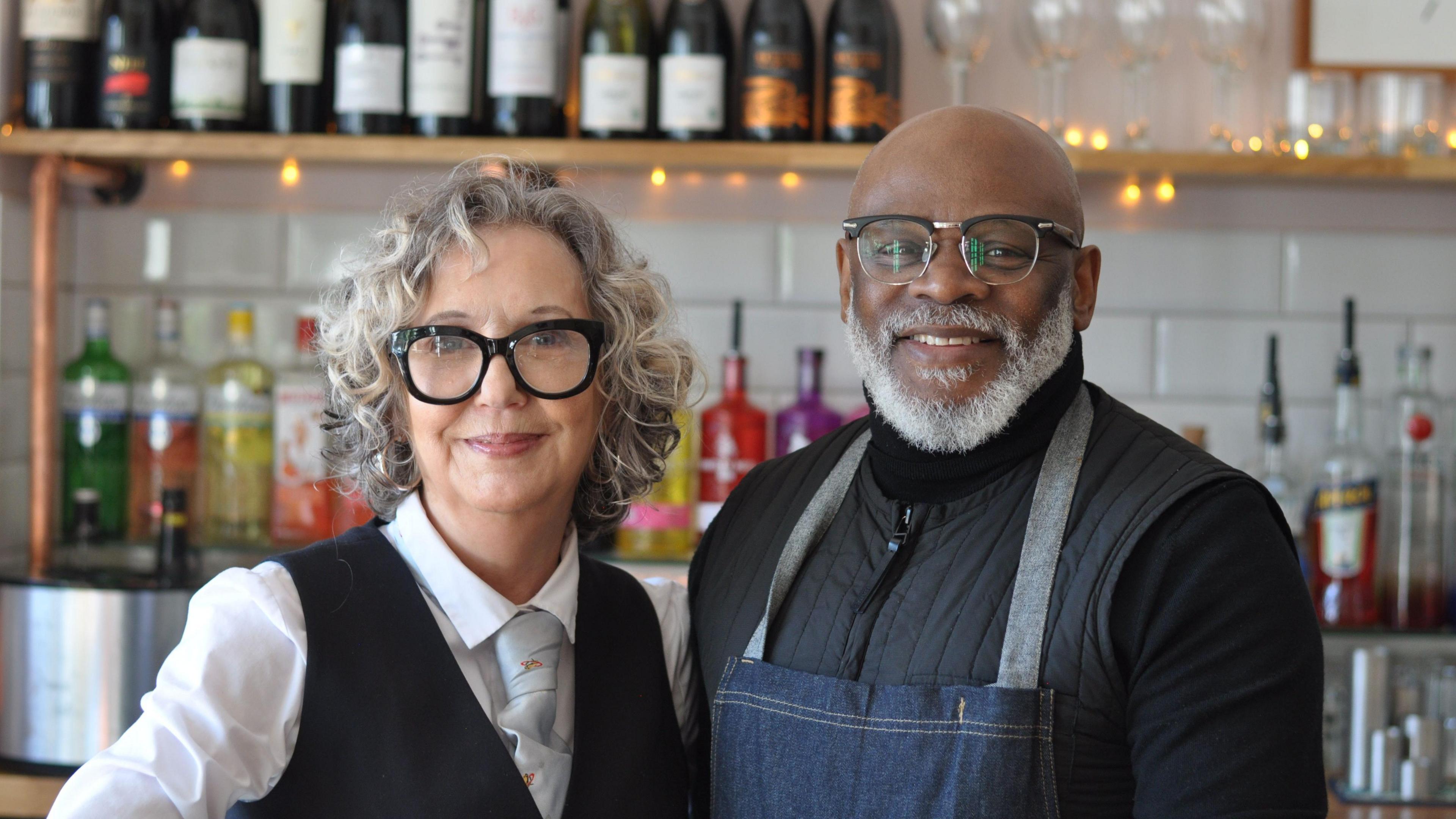 Jayne Casey, with grey curly hair and black rimmed glasses, wears a shirt, tie and black pinafore. Smiling, she stands next to a manwho is also wearing black rimmed glasses. Eric Gooden wears a black jacket and denim apron. They are both standing in front of a bar.