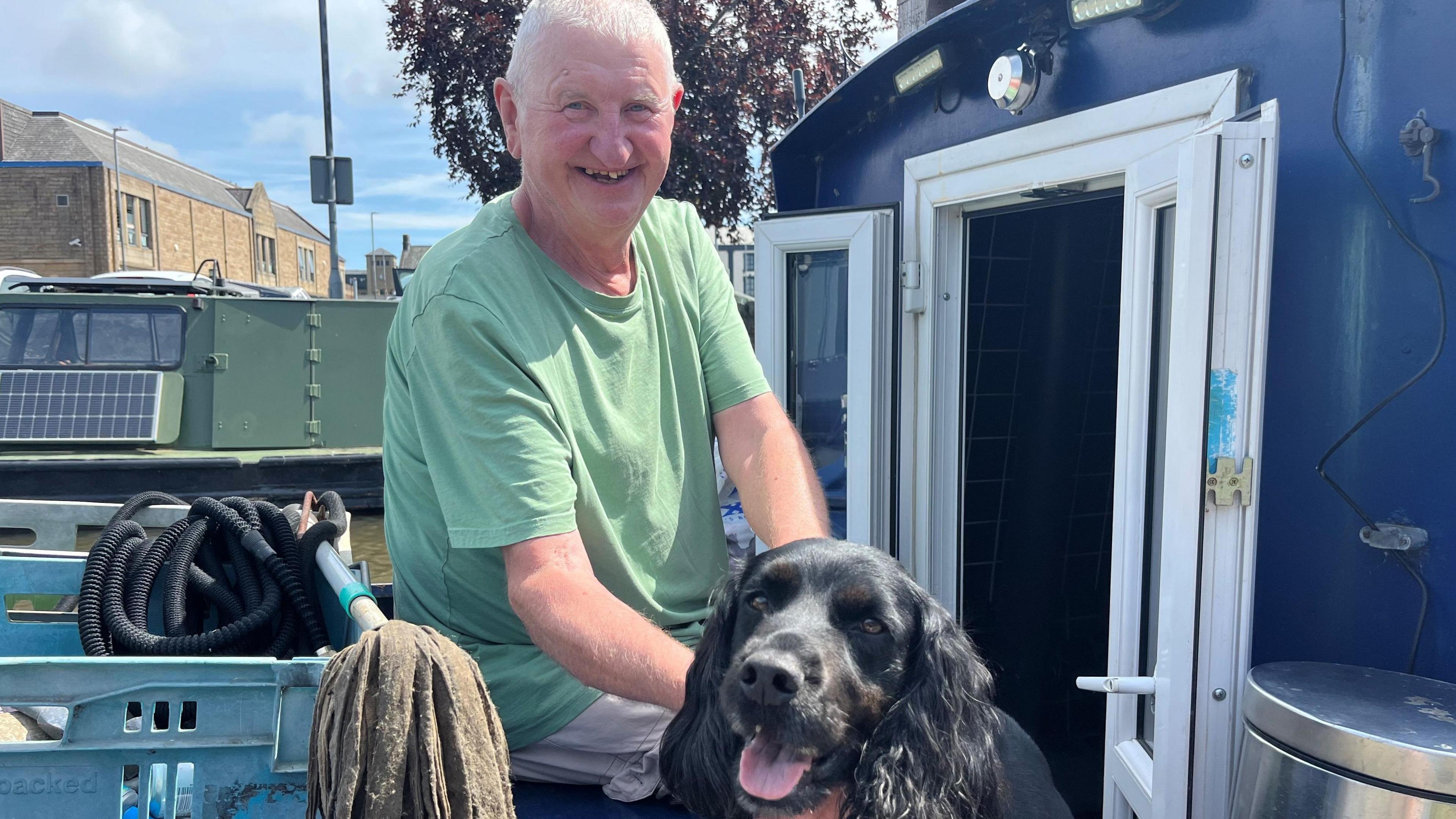 A man in a green T-shirt smiles at the camera. He is petting a spaniel.