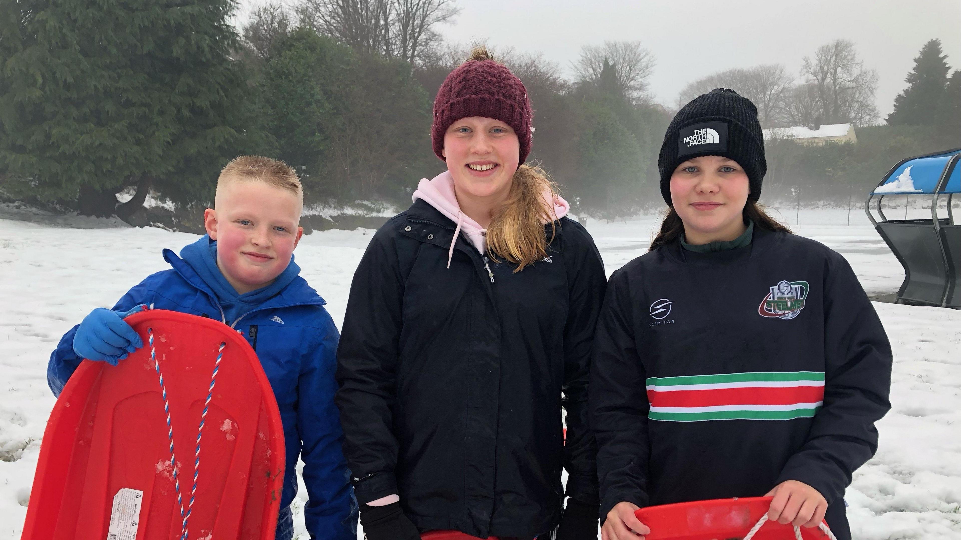 (L-R)A young boy with a blue jacket and gloves on holds his red sledge and smiles at the camera, along with two girls wearing bobble hats and dark zip up rain coats. 