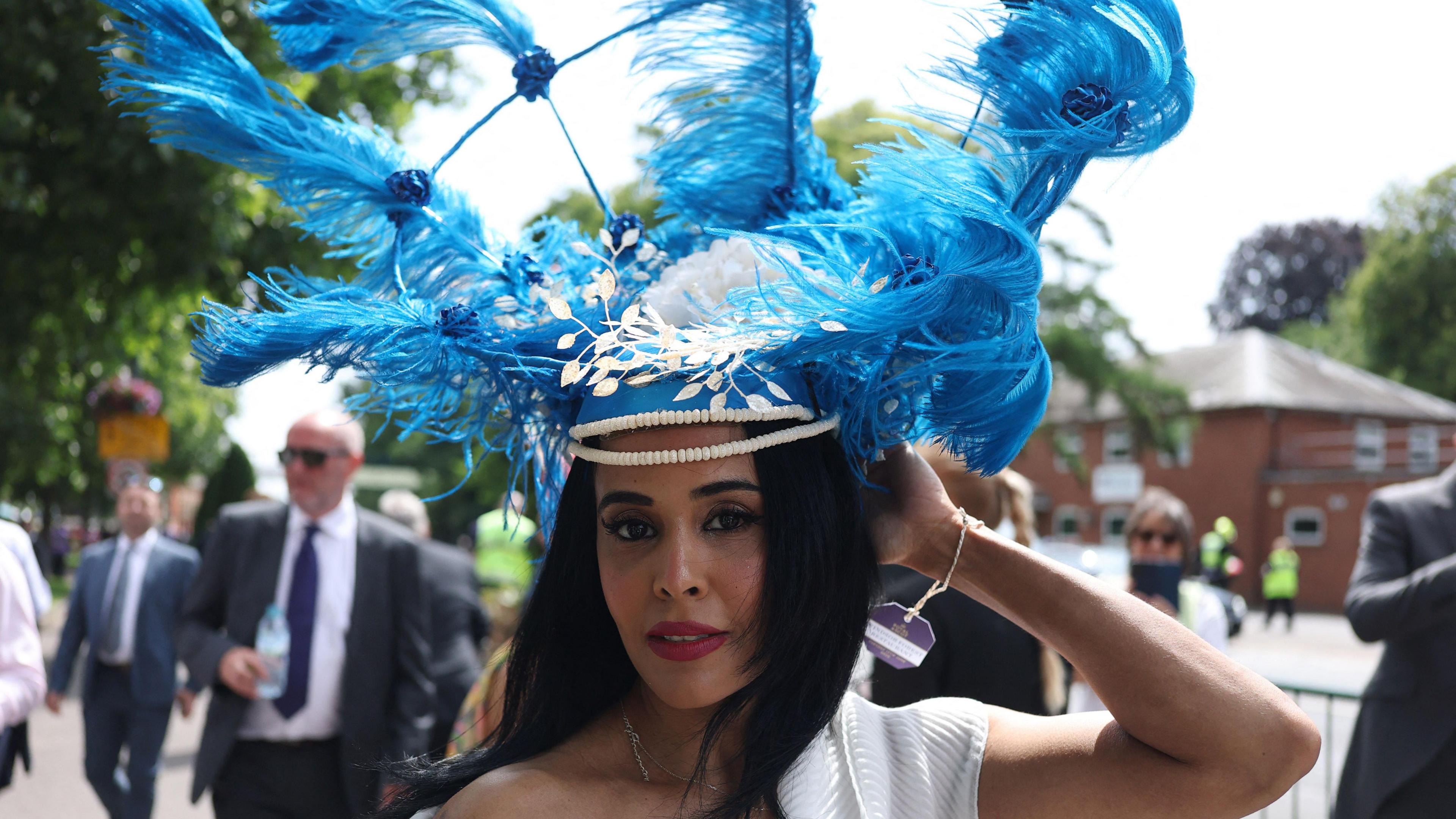 A woman, who is looking at the camera, is wearing a fascinator with blue feathers on it that appear to be blowing in the wind.