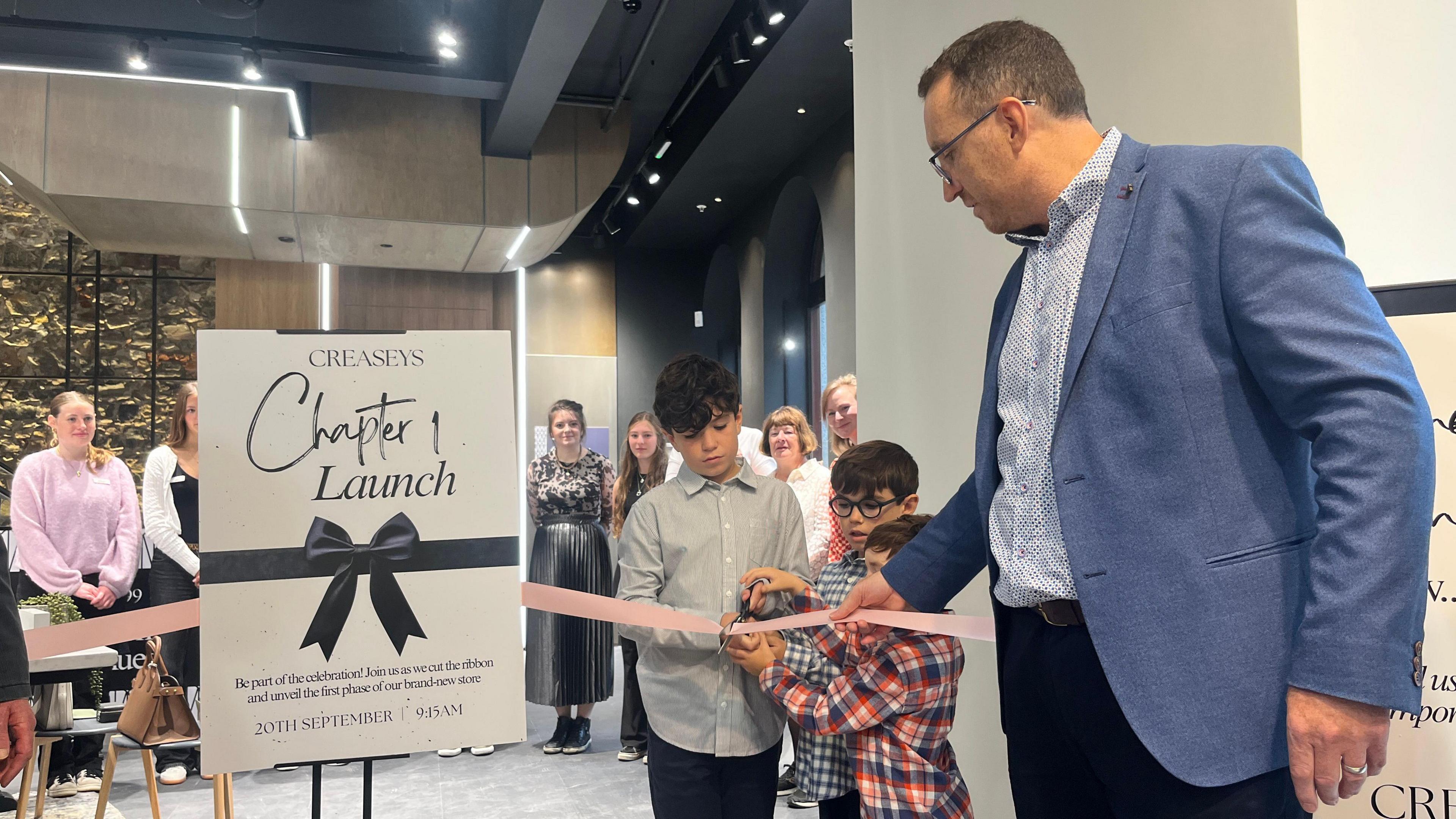 Three small boys cut a ribbon in a shop with a man in a suit - their father - assisting. A sign says Creaseys Chapter 1 Launch - 20 September 2025.