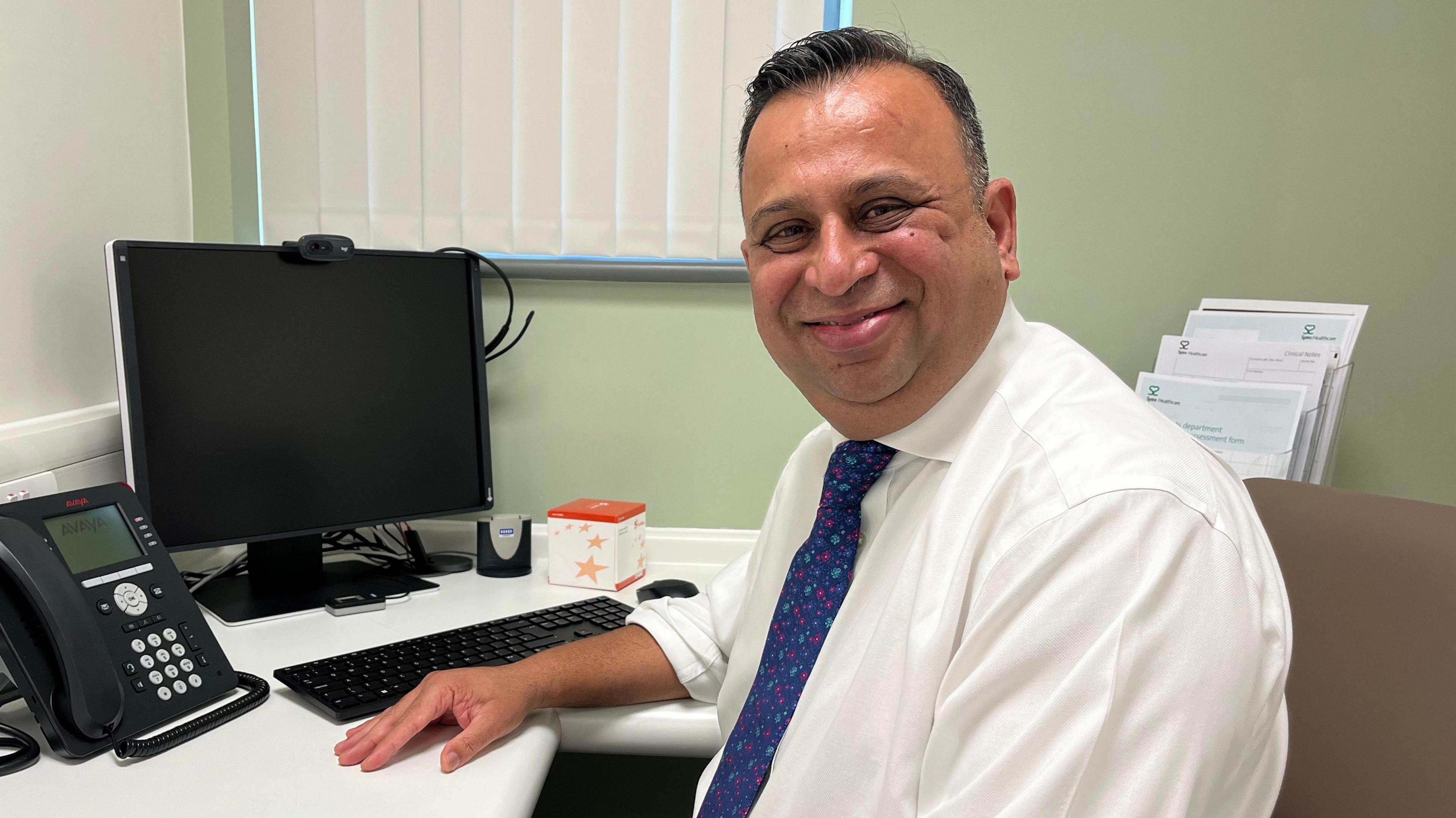 Clinical director for Spire Nayab Haider is sat at a desk. He's wearing a white shirt, blue tie, and smiling